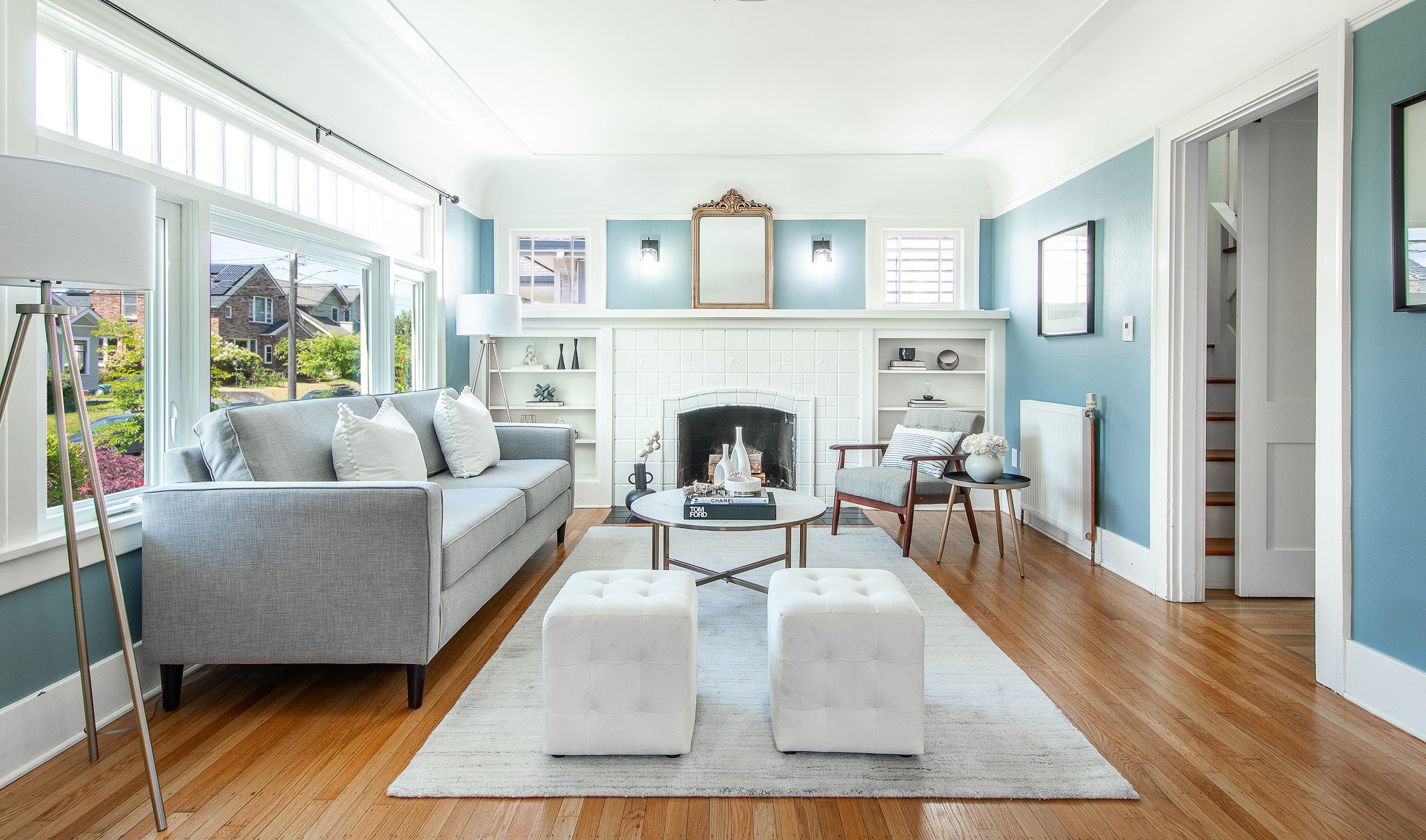 Bright living room with blue walls, white accents, large windows, gray sofa, white ottomans, round coffee table, fireplace, and various decorative items.