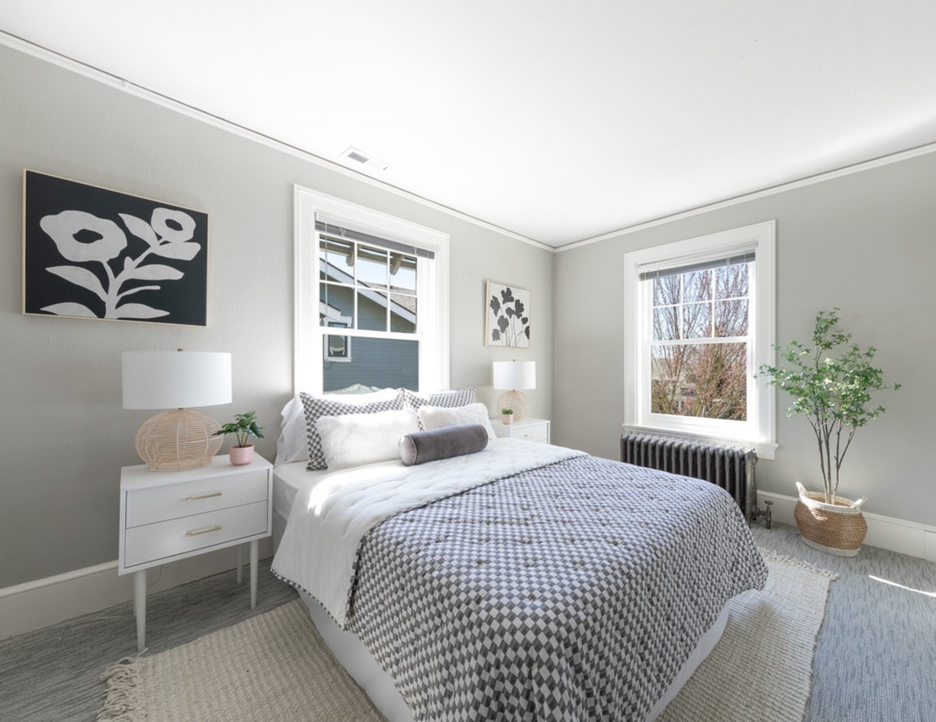 Bright bedroom with two large windows, white bed with black and white checkered bedding, white nightstand with table lamp and small plant, black radiator, light-colored walls with framed black-and-white floral art, and a tall potted plant.