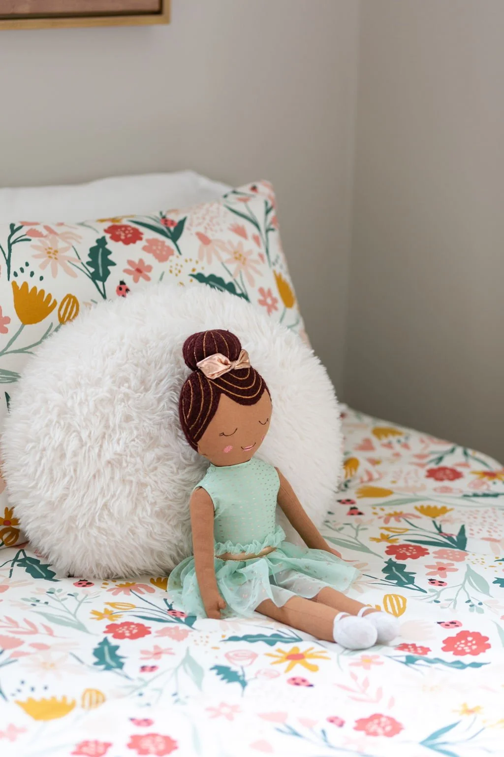 Doll with brown hair in a bun, wearing a light blue dress, sitting on a bed with floral bedding and pillows, surrounded by a white fluffy pillow.