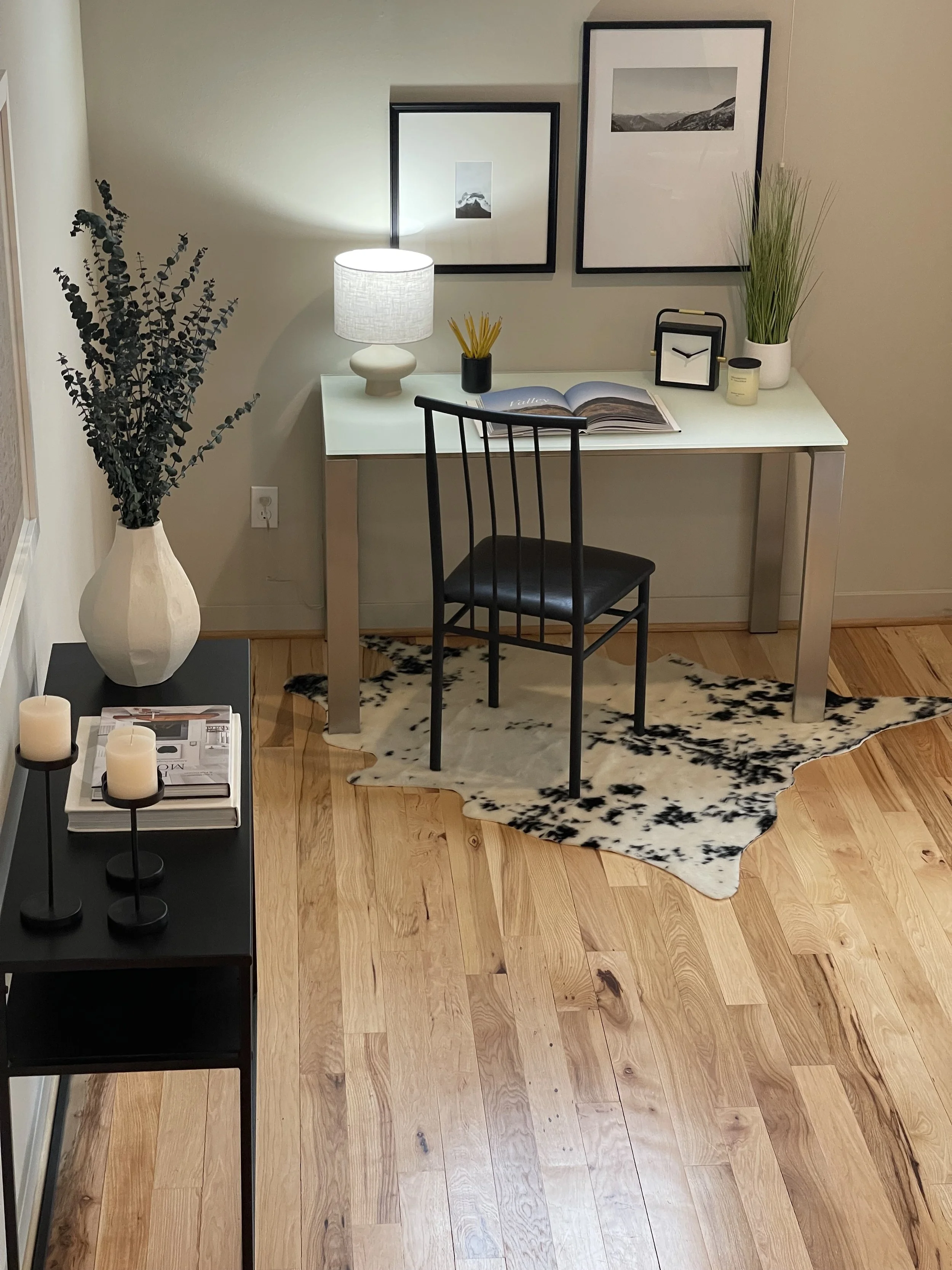 A home office workspace with a white desk, black chair, and decorative items, including framed black-and-white landscape photos, a table lamp, a green potted plant, and a cowhide rug on a wooden floor.