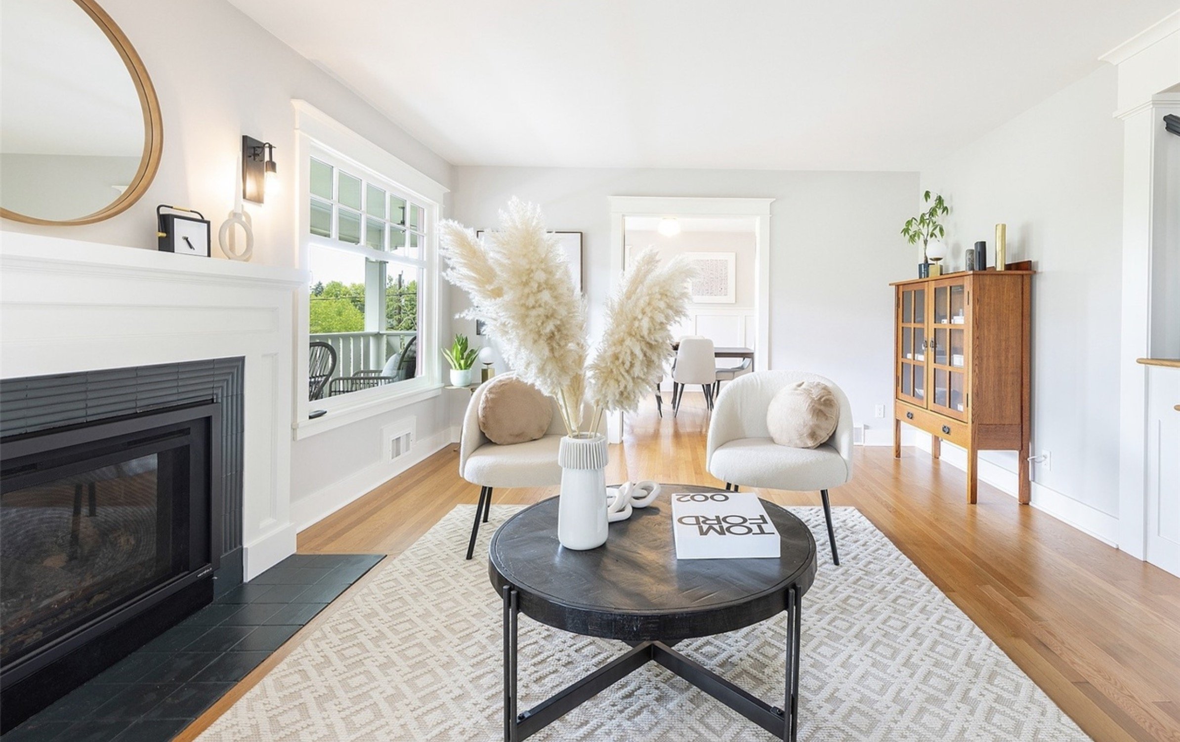 A bright living room with white walls, a large window, a black fireplace, and a wooden cabinet. There are two cream-colored chairs with pillows, a black round coffee table with decorative objects, and a beige patterned rug. A dining area with a table