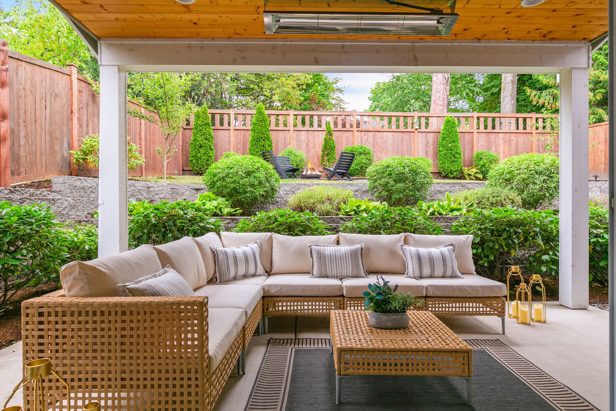 Outdoor patio with beige sectional sofa, striped pillows, woven coffee table, surrounded by greenery, trees, and a wooden fence.