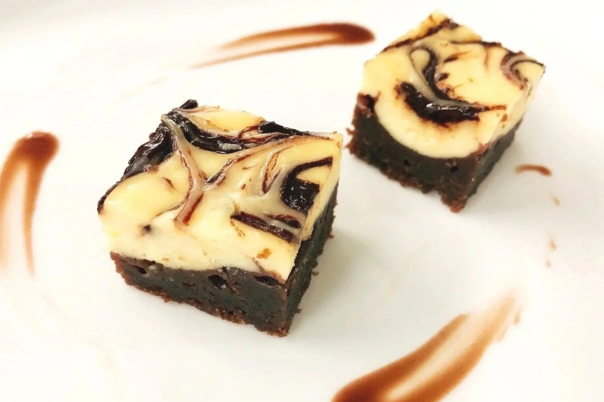 Two small square pieces of cheesecake with a fudge base, topped with swirled cream cheese mixture, on a white plate.