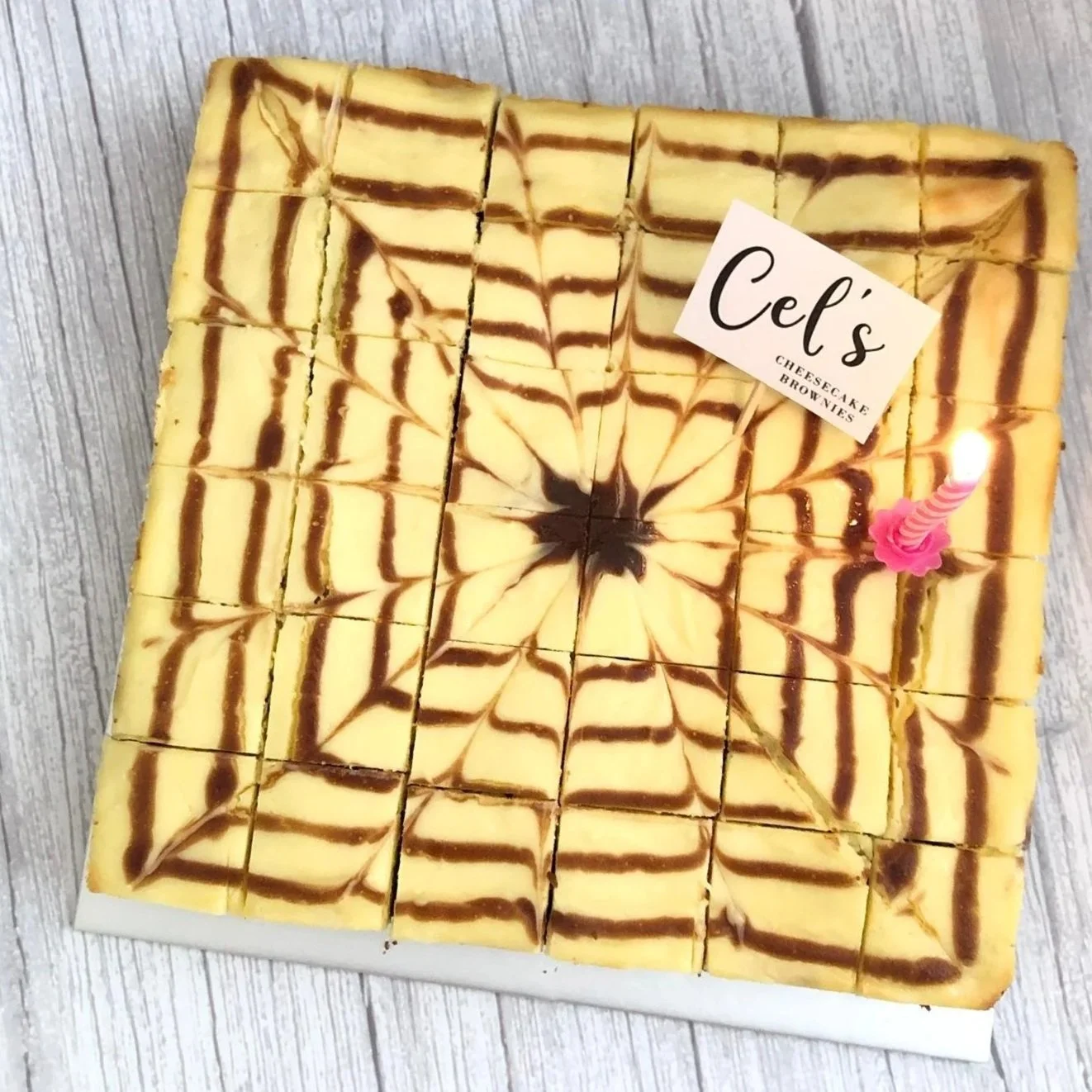 A square tray of cheesecake brownies with a marbled swirl pattern, sliced into small square pieces, and a pink birthday candle with a pink holder on the right side.