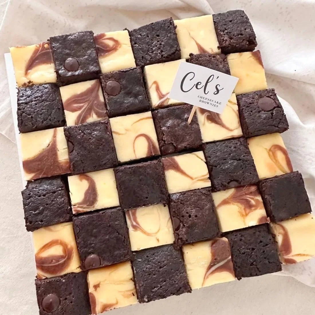 A checkerboard tray of mixed cheesecake and brownie squares, with some topped with chocolate chips, labeled "Cef's Cheesecake Brownies."
