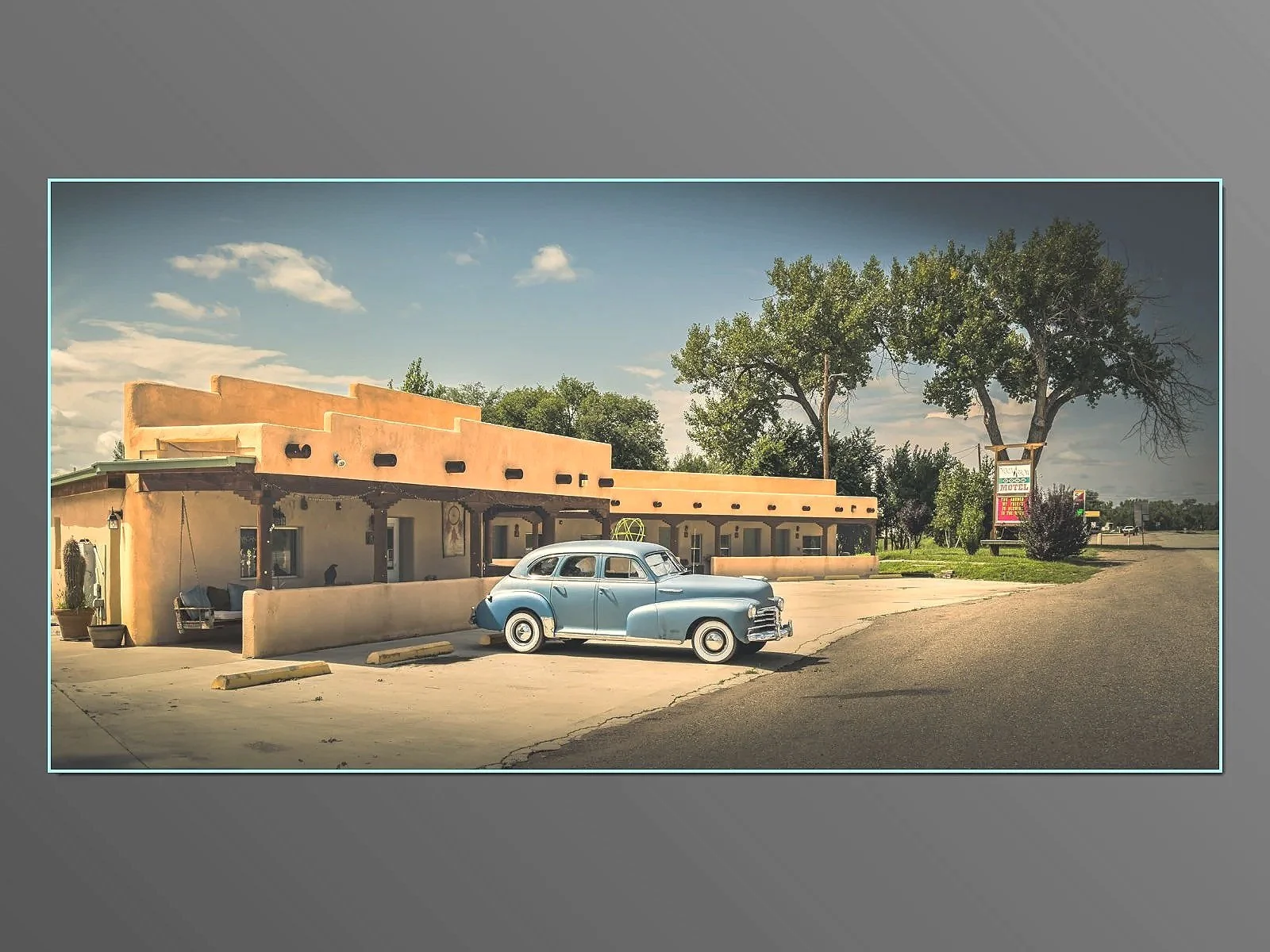 A vintage blue car parked in front of a southwestern style motel with a sign, large trees, and a clear sky in the background. Springer NM.
