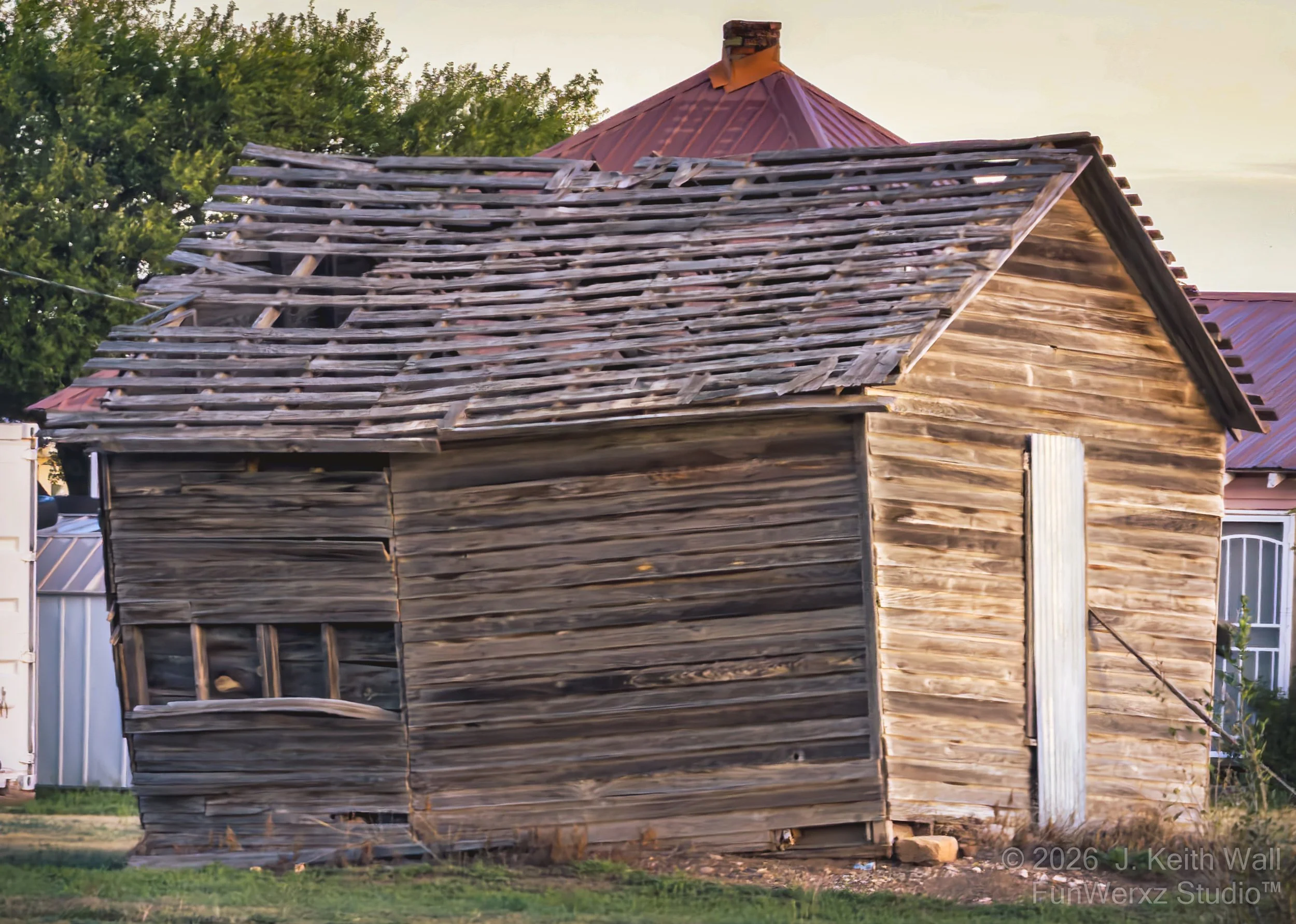 An old, weathered wooden house with a damaged roof, missing many shingles, in a residential neighborhood with trees and other buildings visible in the background.