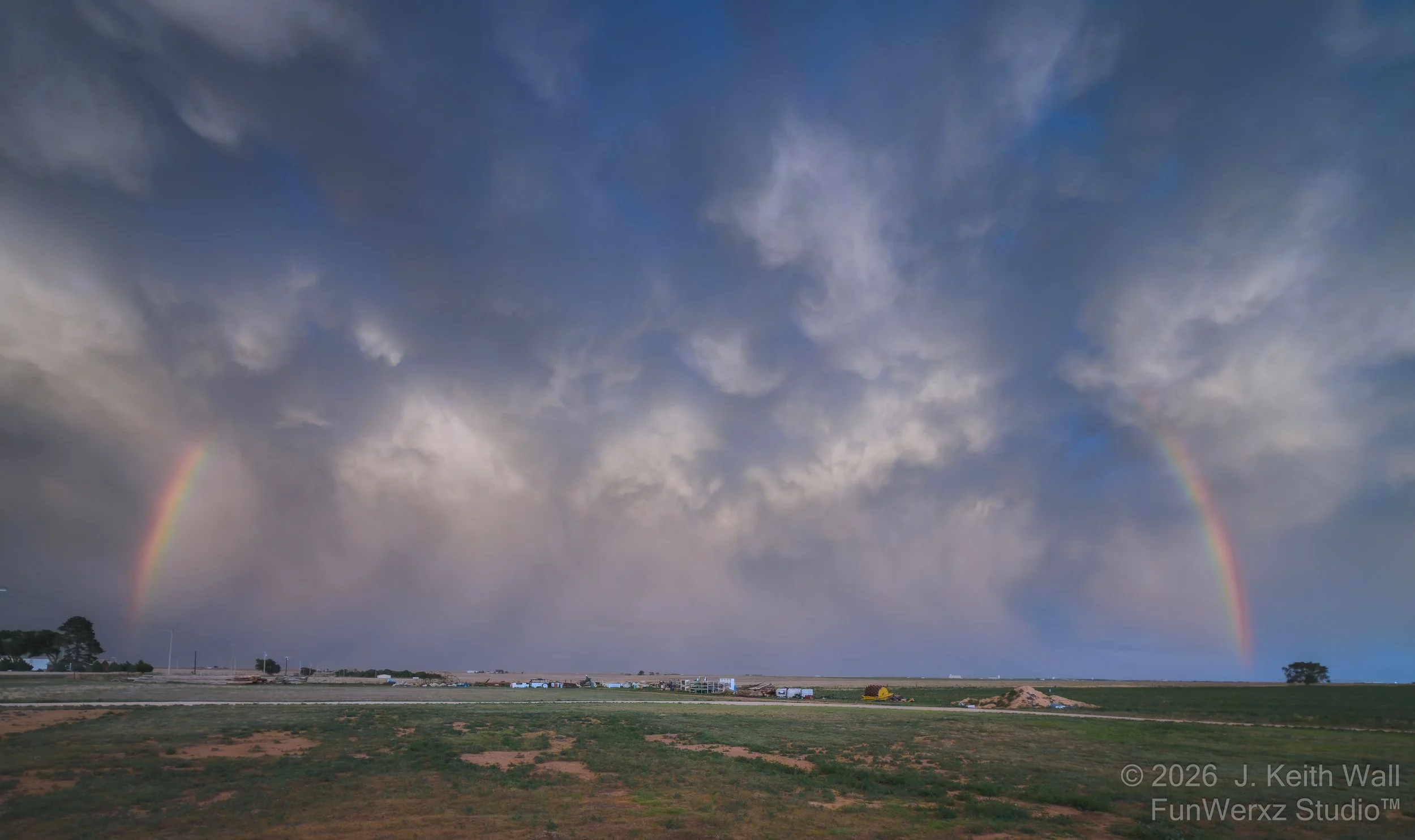 A landscape view beneath stormy clouds with a rainbow appearing in the sky, spanning from the left to the right horizon.