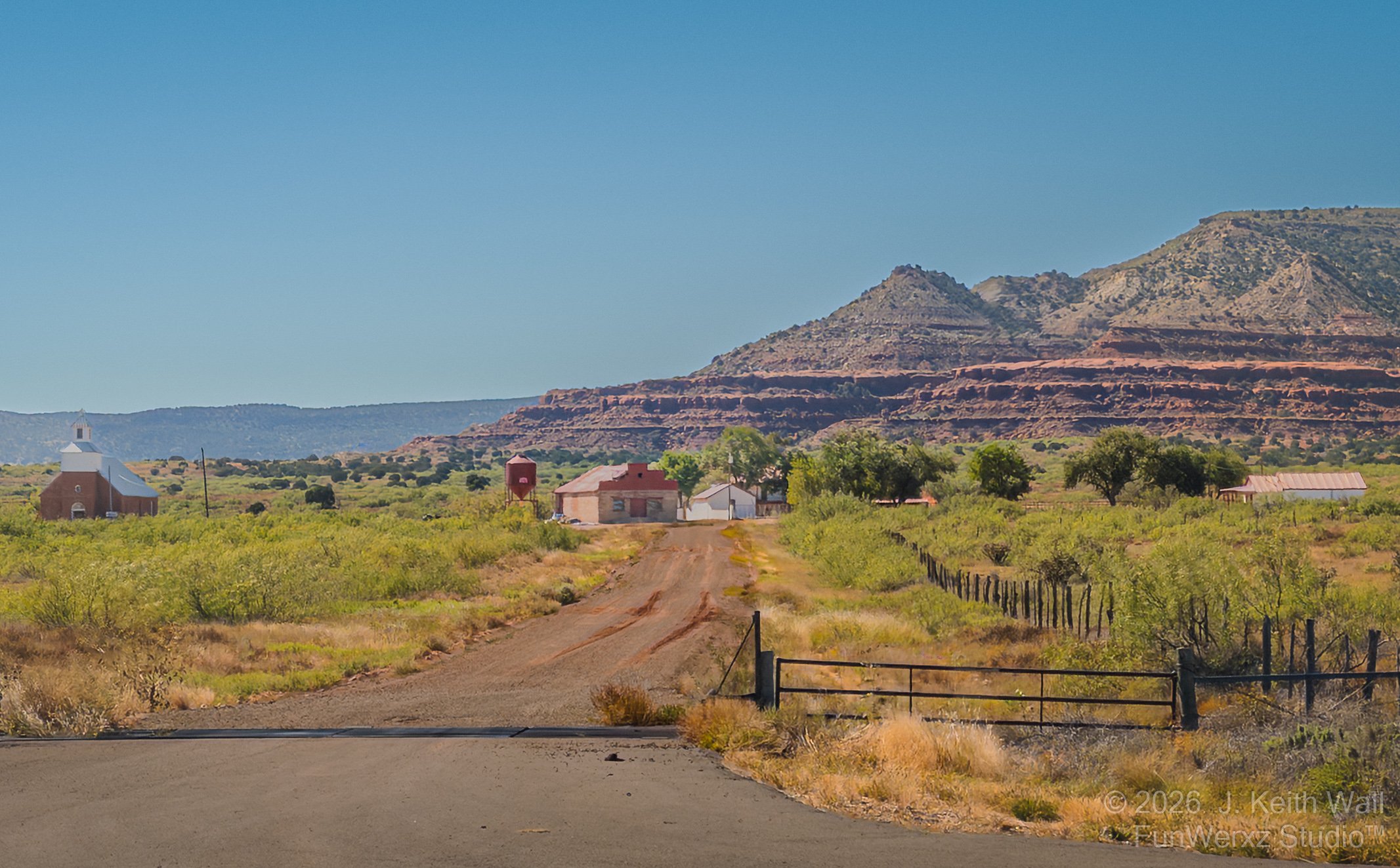A dirt road leading to a small rural farmhouse and barn, with a church on the left, green shrubbery, and mountains in the background under a clear blue sky.