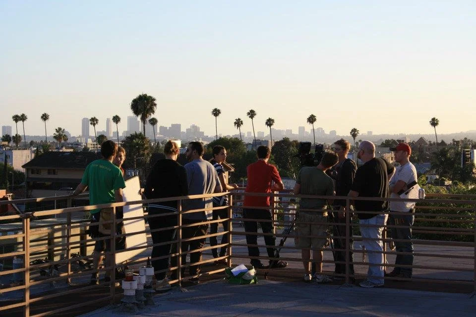 Group of people on a rooftop observing cityscape with palm trees and skyline in the background at sunset.