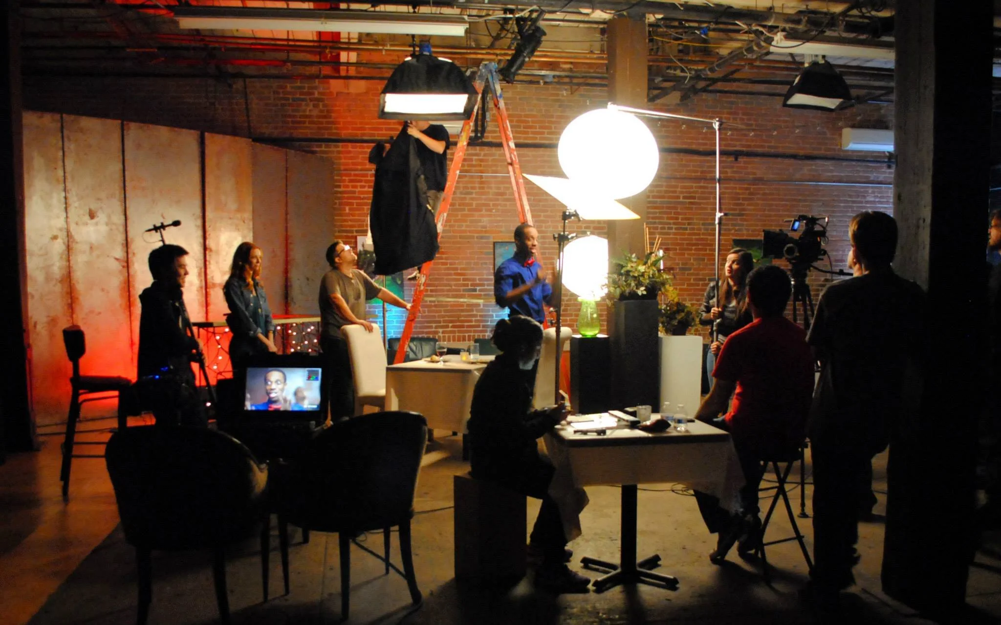 Indoor film or video production set with crew members, lighting equipment, and a brick wall background.