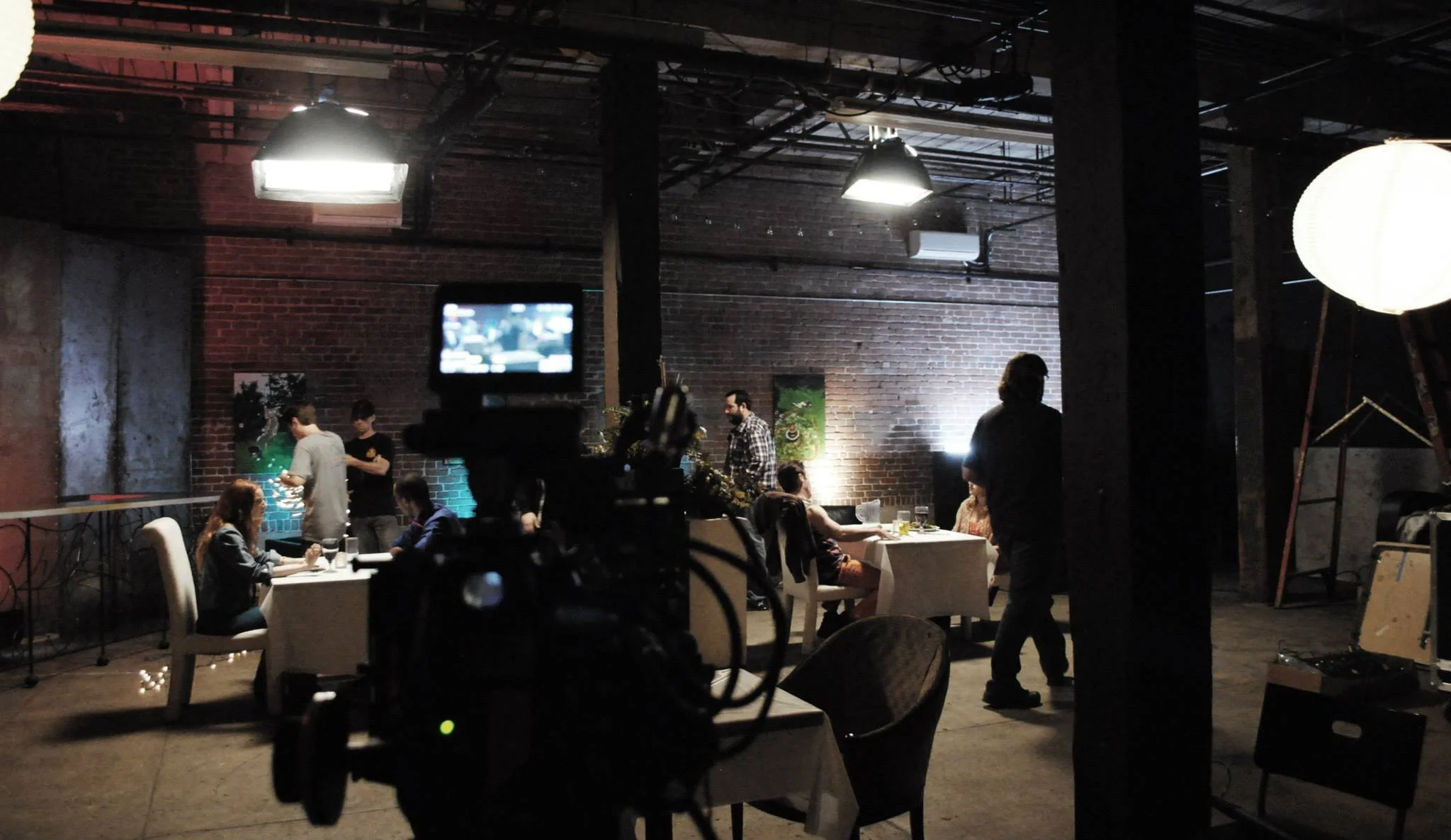 A restaurant or event space with brick walls and industrial ceiling lighting. Several tables are occupied by people dining, with some staff serving. A camera is positioned in the foreground.