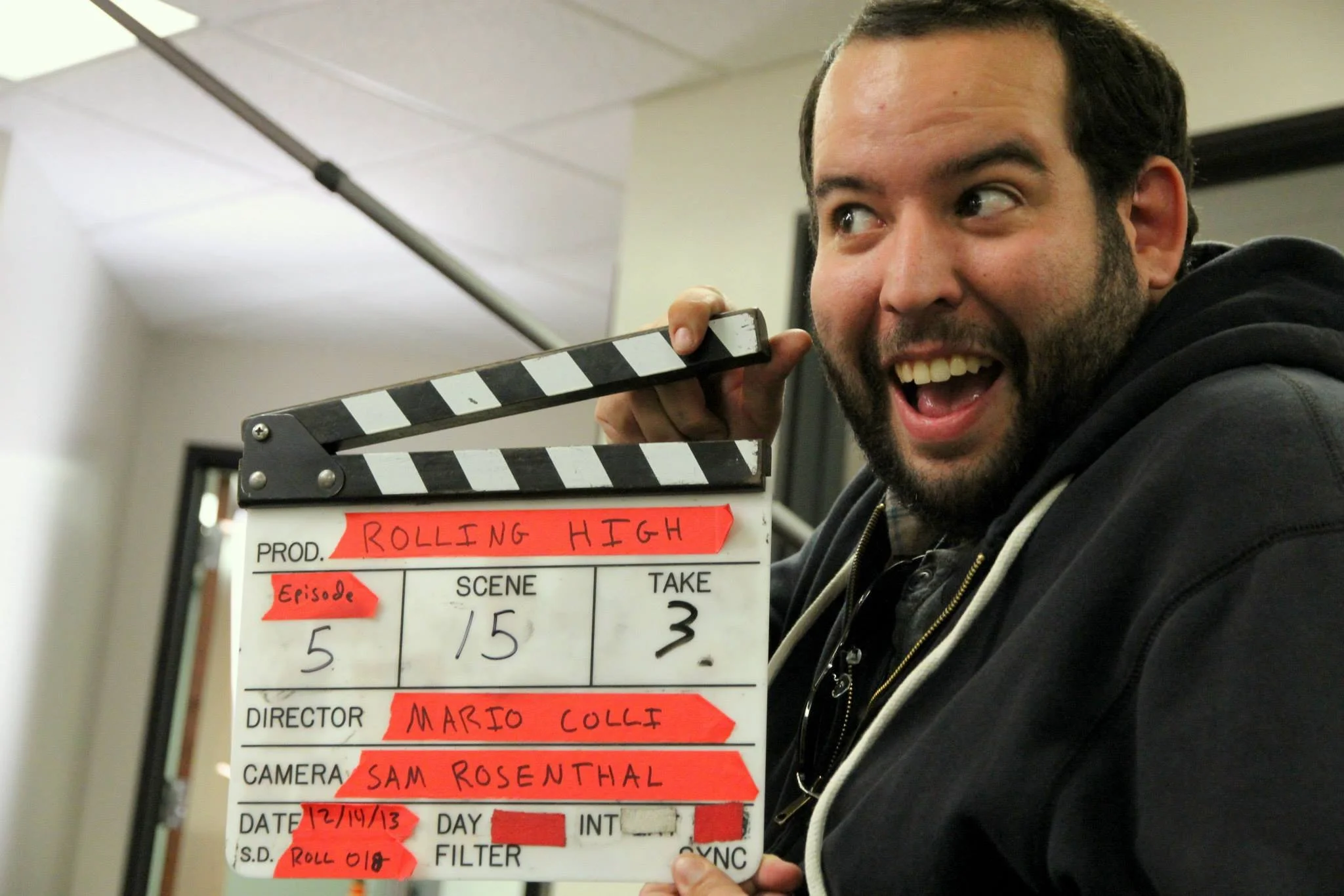 A man with a beard and dark hair smiling and holding a film slate labeled 'Rolling High' in a room with a white ceiling and walls.