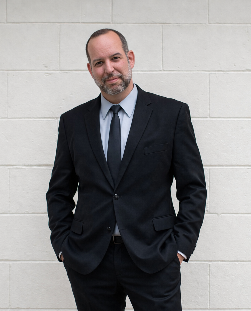 A middle-aged man with a beard and receding hairline, wearing a black suit, light blue shirt, and dark tie, stands against a light-colored brick wall with his hands in his pockets, looking at the camera with a slight smile.