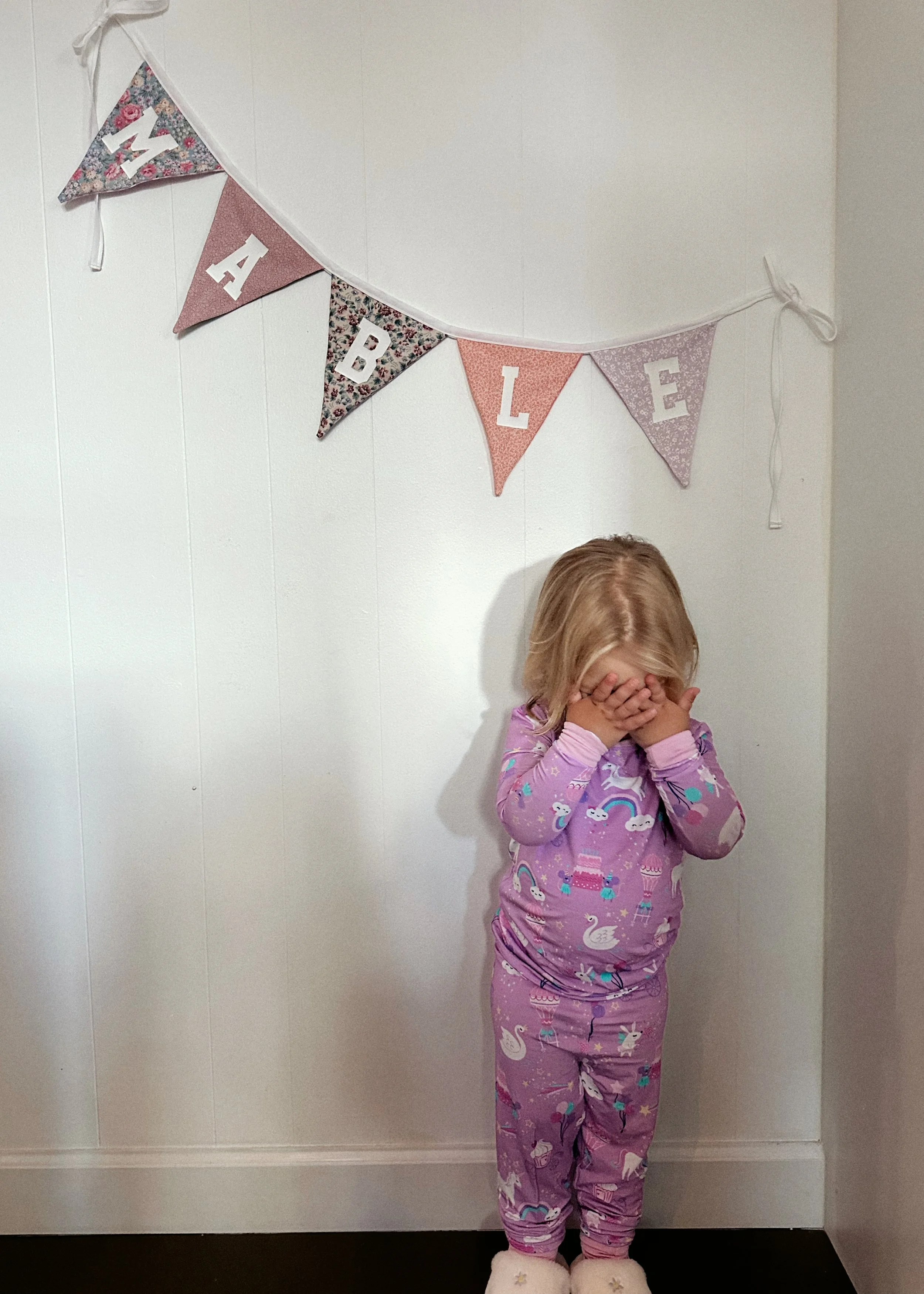 A young girl with blonde hair in pajamas with unicorn, rainbow, and swan patterns standing against a wall, covering her face with her hands, wearing fluffy slippers with star designs, and a decorative bunting with floral and pink fabric triangles reading "HAPPY DAY" hanging above her.