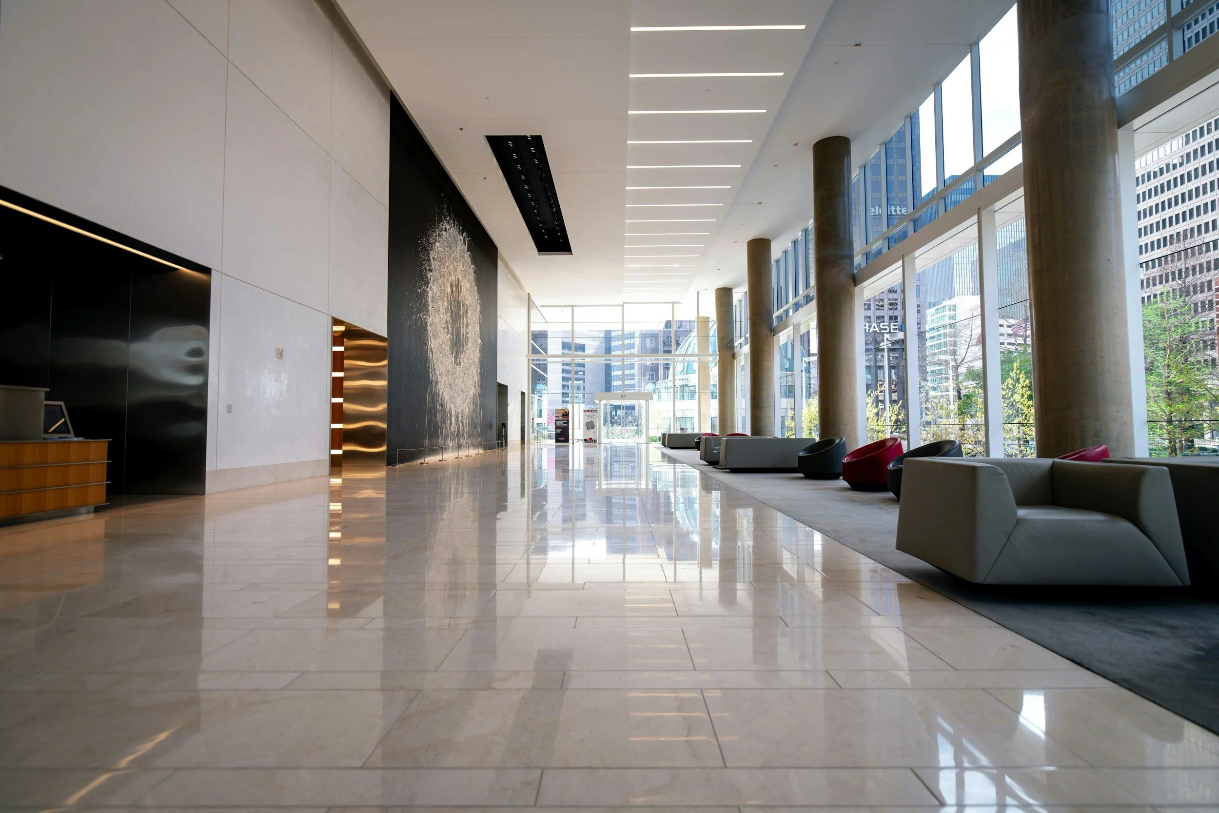 Modern hotel lobby with large floor-to-ceiling windows, white marble floors, scattered contemporary chairs, and an indoor water feature wall with water cascading down.