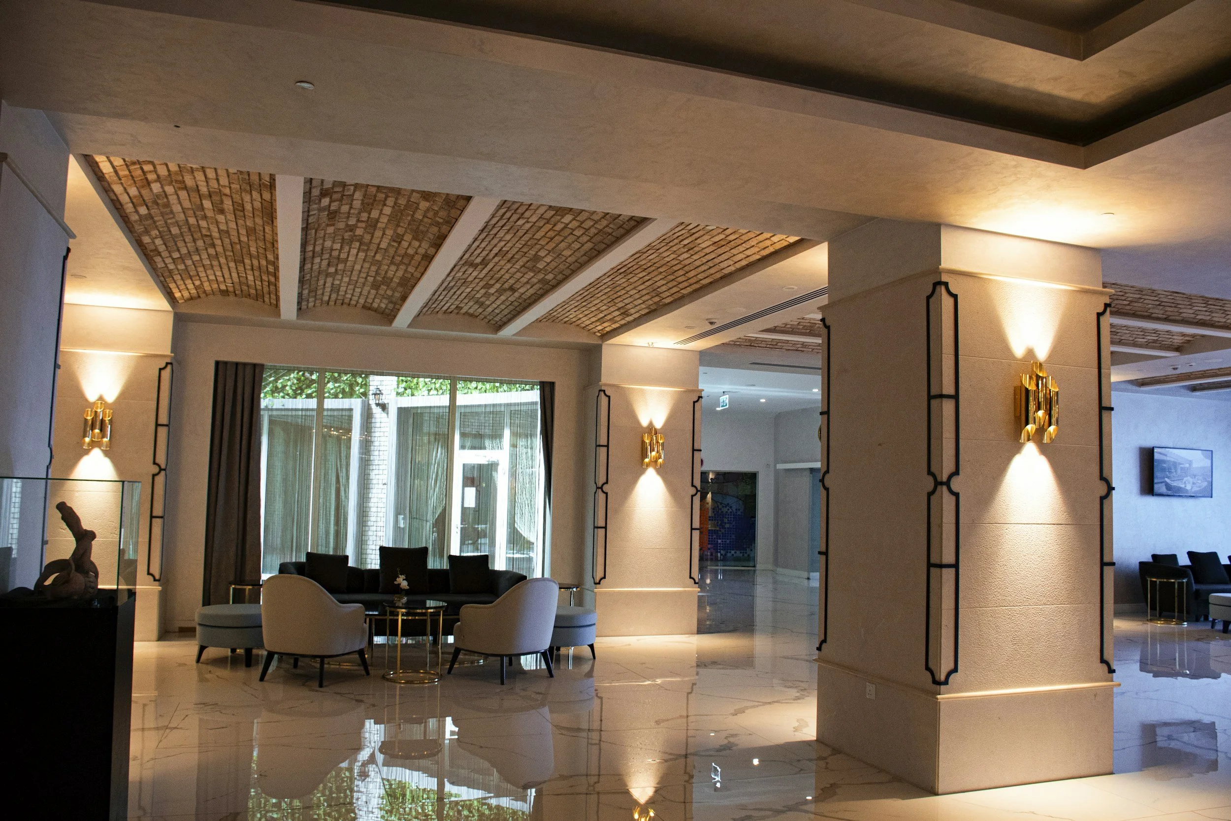 Interior of a modern hotel lobby with a high ceiling featuring brick arches, large windows, and elegant lighting fixtures. There are chairs and a small table in the seating area, with decorative gold accents.
