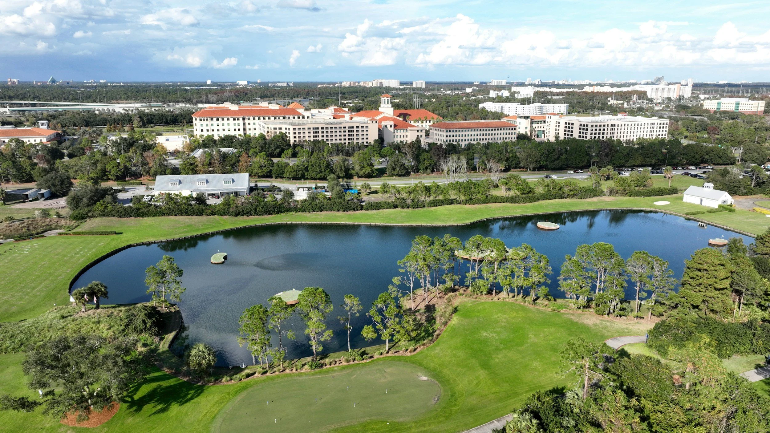 Aerial view of a large complex with multiple beige buildings with red roofs, surrounded by lush greenery and a pond with trees, and a road. In the background, there are more buildings and a highway.
