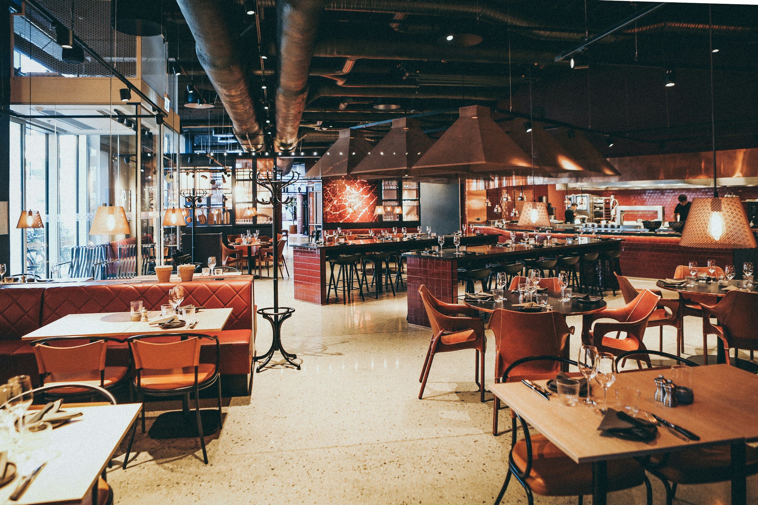 An interior view of a modern restaurant with tables set for dining, warm lighting, and decorative elements including large metal hood vents and a red brick wall with lighting fixtures.
