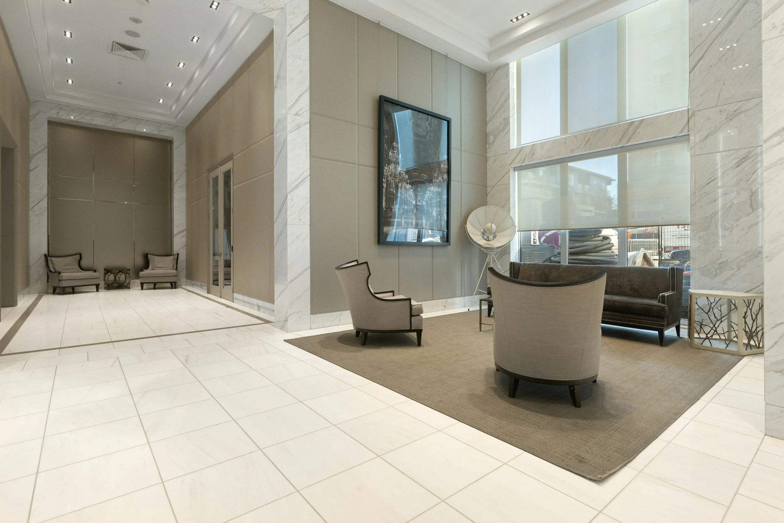 Modern lobby with white tiled floors and light-colored walls, contemporary chairs, large windows with shades, a mounted flat-screen TV, and decorative elements.
