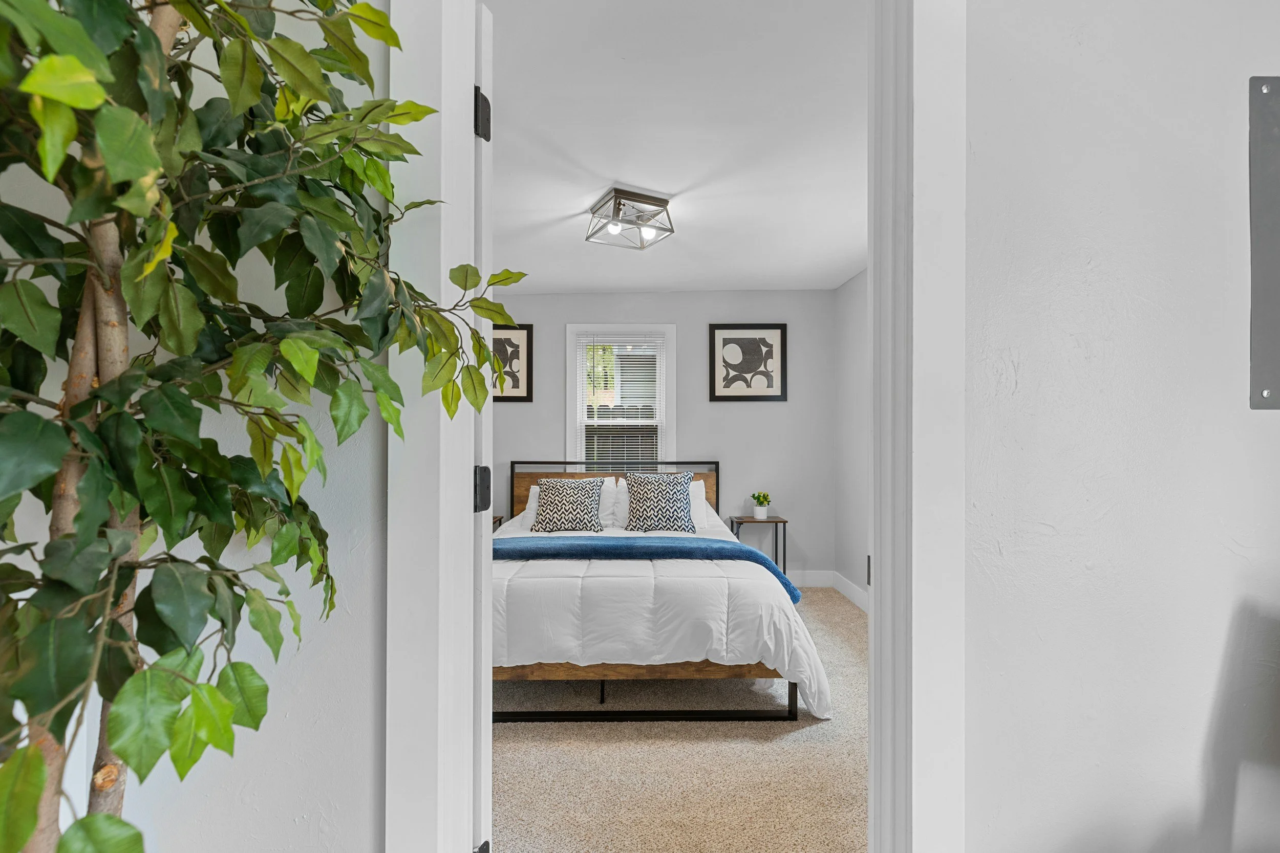 A view into a bedroom through a doorway, with a bed, two side tables, framed wall art, a window with blinds, and a ceiling light fixture, with a leafy plant partially visible on the left.