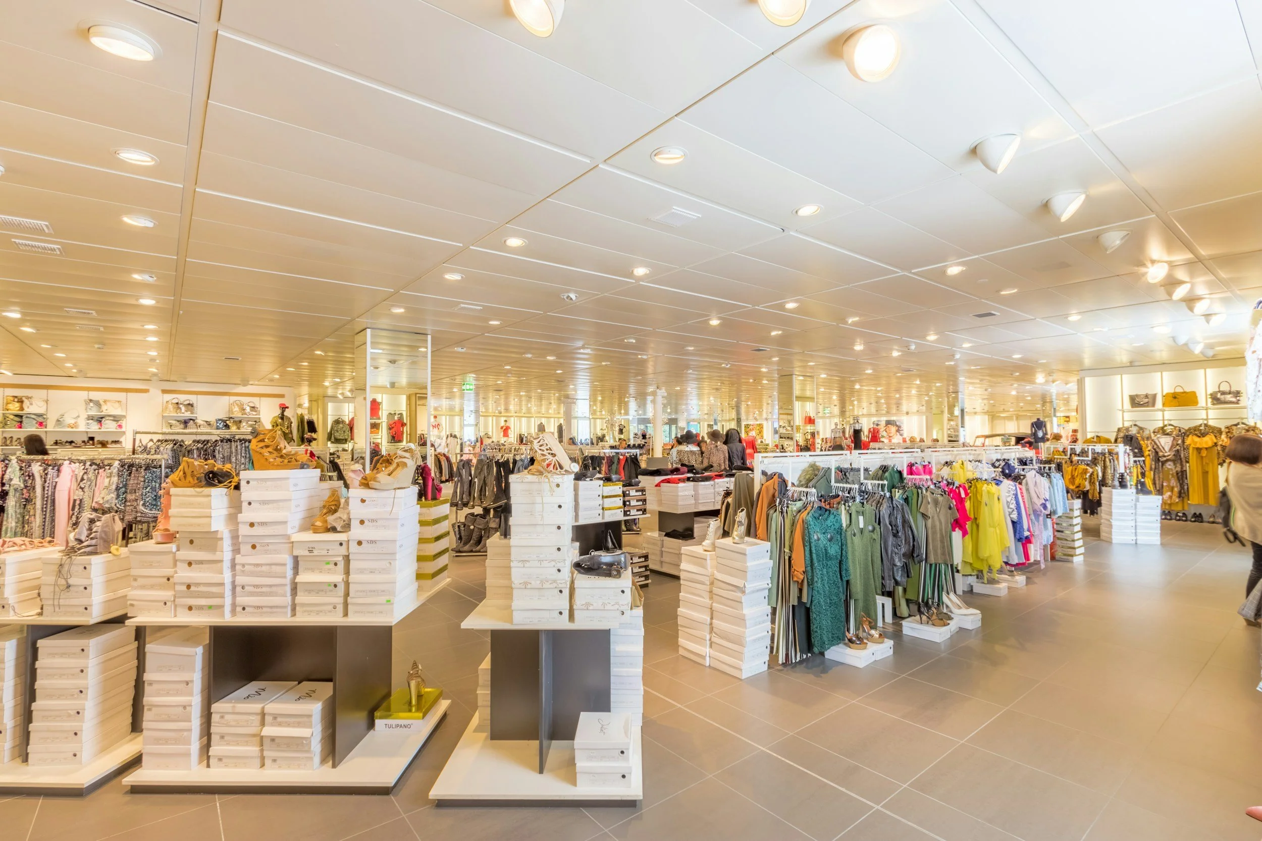 Clothing section inside a department store with racks of dresses, shoes displayed on tables, and shoppers shopping.