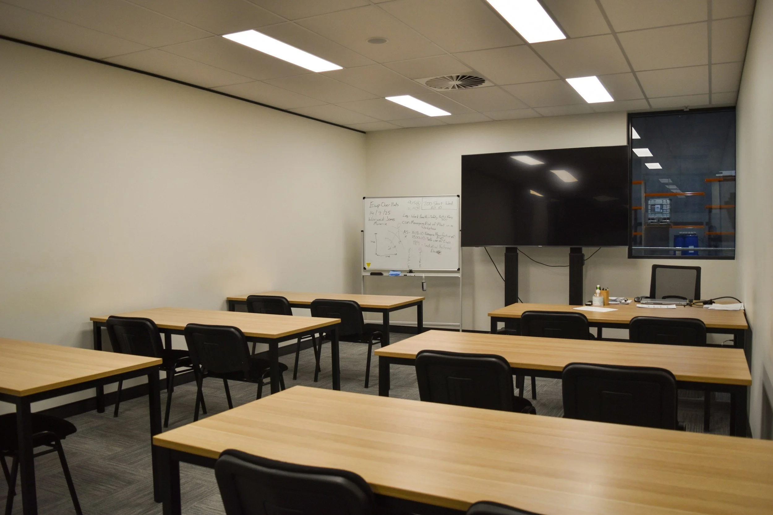 An empty conference room with tan tables and black chairs, a whiteboard with writing, a large screen, and a window showing a storage room with shelves and a trash can.