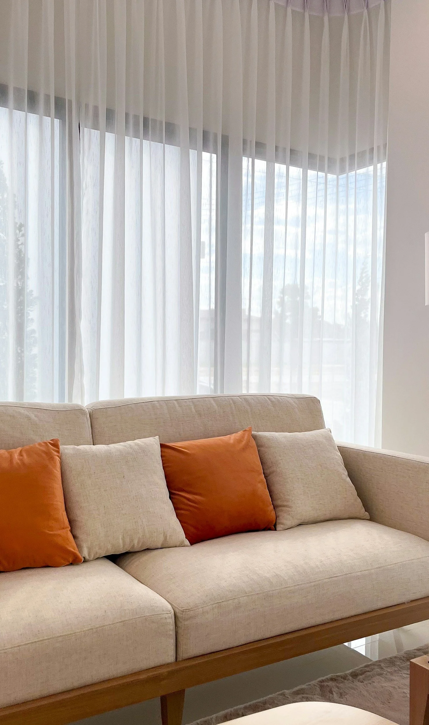 A beige sofa with orange and beige cushions positioned in front of a large window with sheer white curtains. The window shows some outdoors with a bright, possibly cloudy sky.