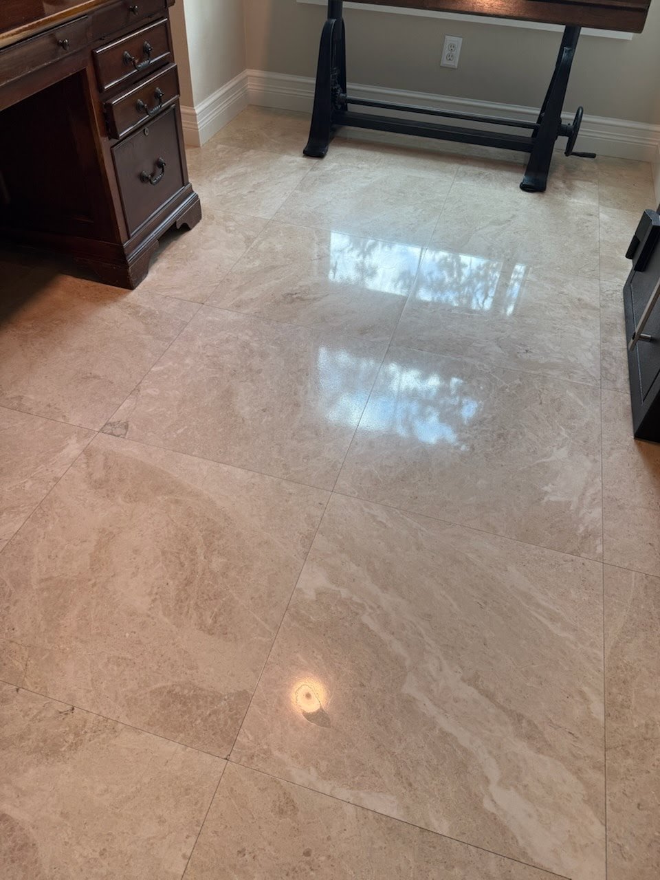 A shiny, polished tile floor in a room with a dark wooden cabinet and a black tabletop stand, with a wall outlet and baseboard in the background.