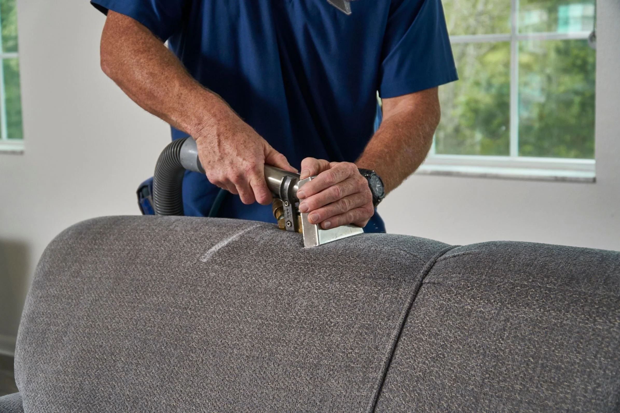 A person in a blue shirt using a vacuum cleaner on a gray upholstered piece of furniture in a room with large windows.