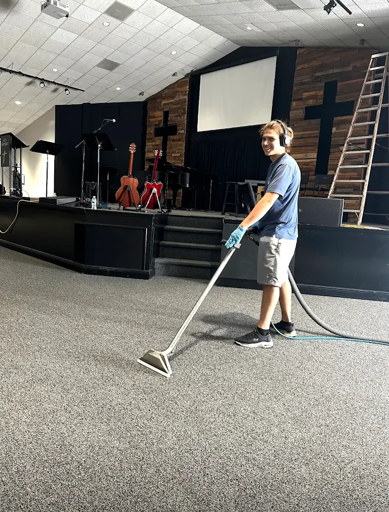 A young man wearing headphones, gloves, a blue t-shirt, and shorts vacuums a carpeted floor inside a church or auditorium with a stage, musical instruments, and a ladder in the background.
