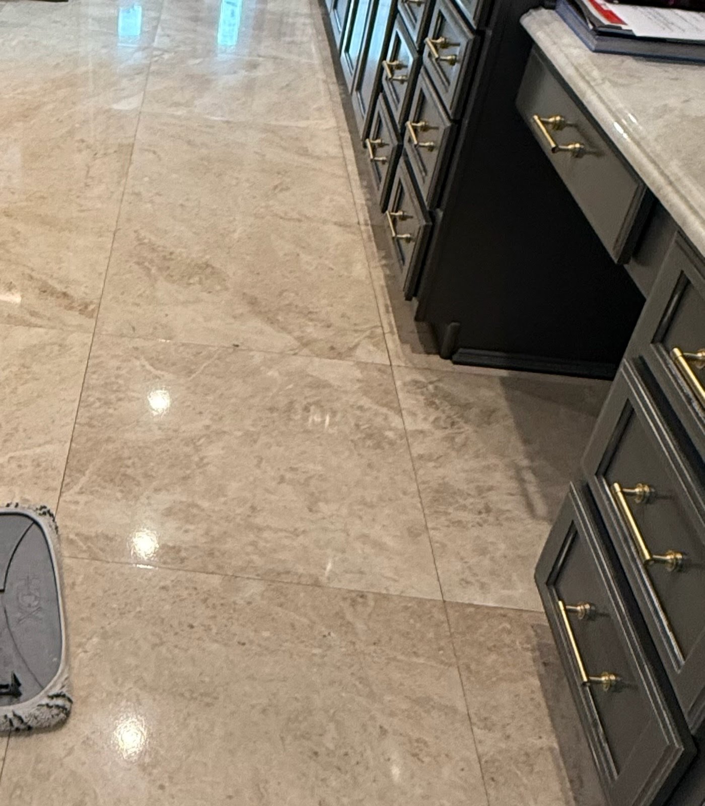 A kitchen counter with dark cabinets and gold handles, beige marble floor tiles, a small part of a gray appliance visible in the lower left corner, and a few magazines or books placed on the counter.