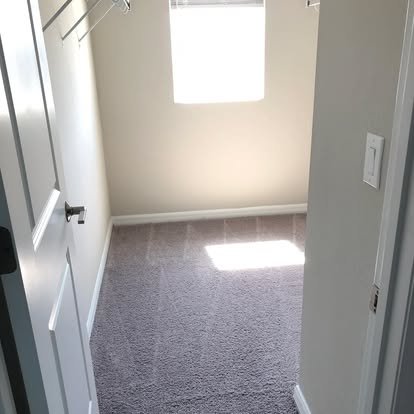 Empty small room with beige walls, gray carpet, a window with closed blinds, and a light switch on the wall.