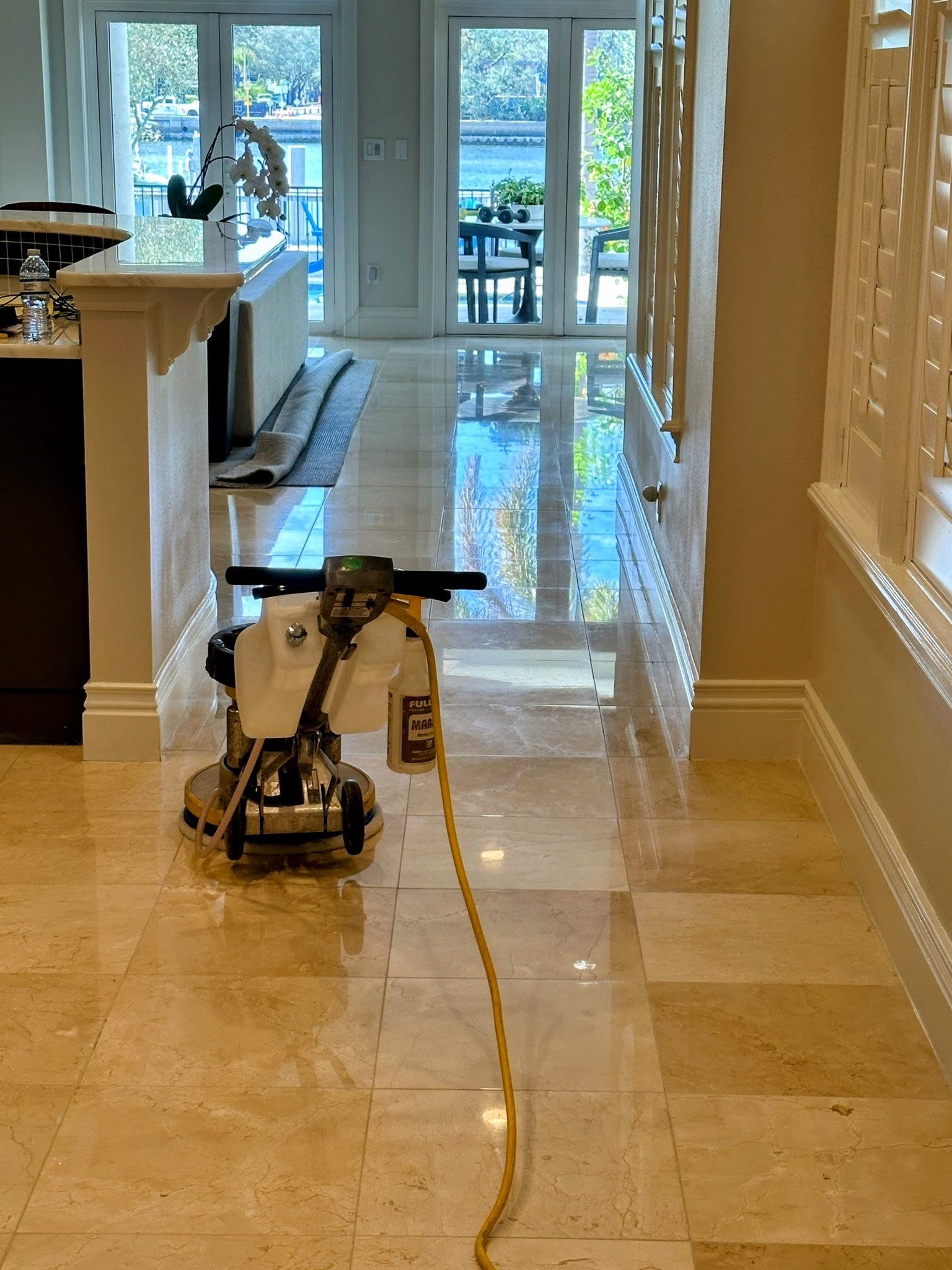 View of a hallway with beige tile floors, a yellow extension cord on the floor, a child's tricycle, and a view of a patio through glass doors with a table and chairs outside.