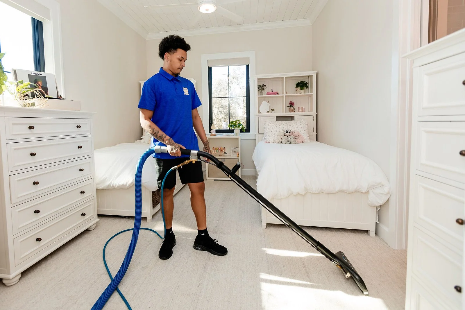 A person vacuuming a white bedroom with two beds, white dressers, shelves, and a window with sunlight.