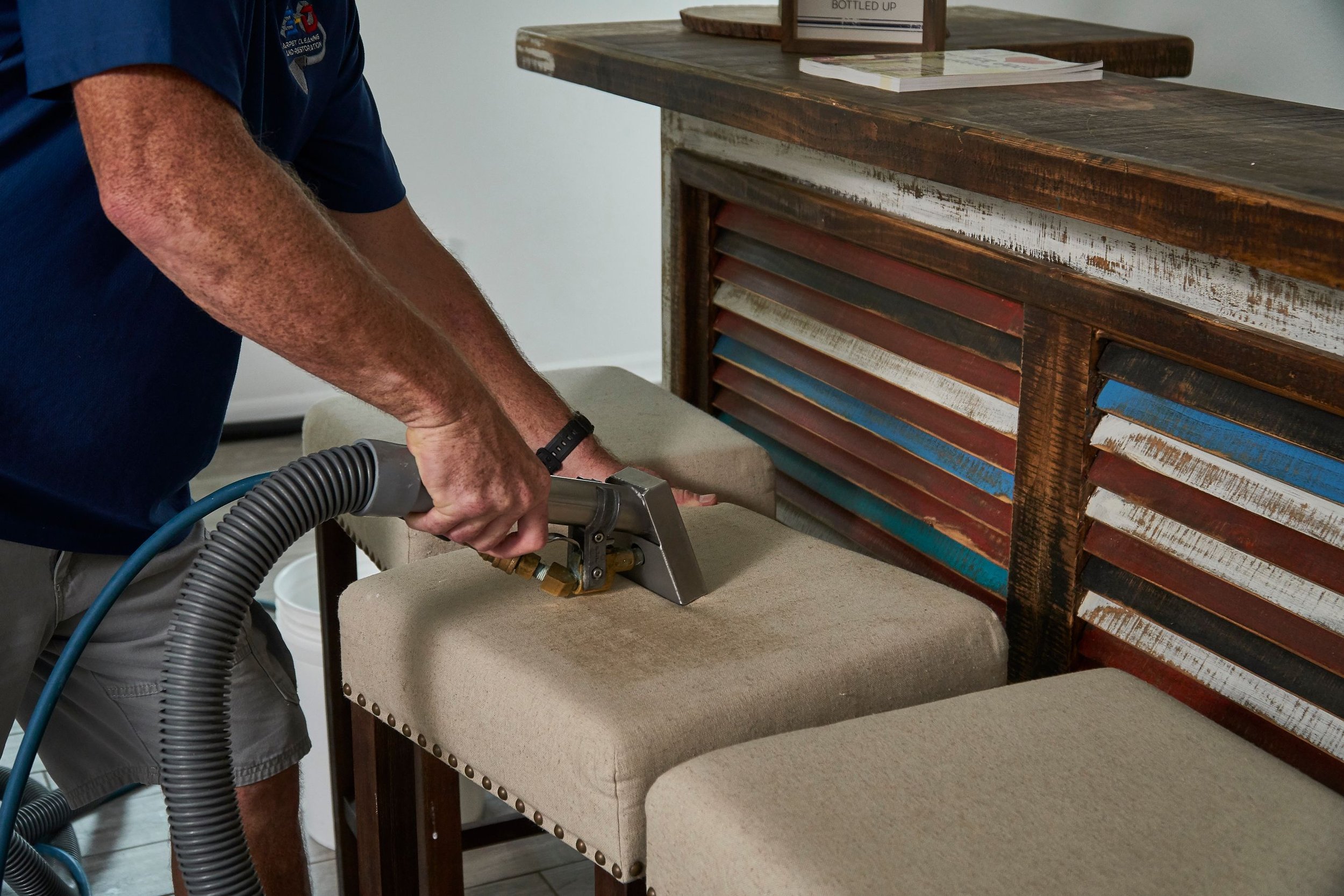 A person using a vacuum cleaner on a beige upholstered chair with a wooden table nearby. The table has a stack of magazines and a framed sign that says 'BOTTLED UP'. The chair is against a wall with some tile flooring visible.