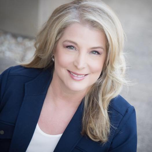 A smiling woman with blonde hair wearing a navy blazer and white top, sitting indoors with a neutral background.