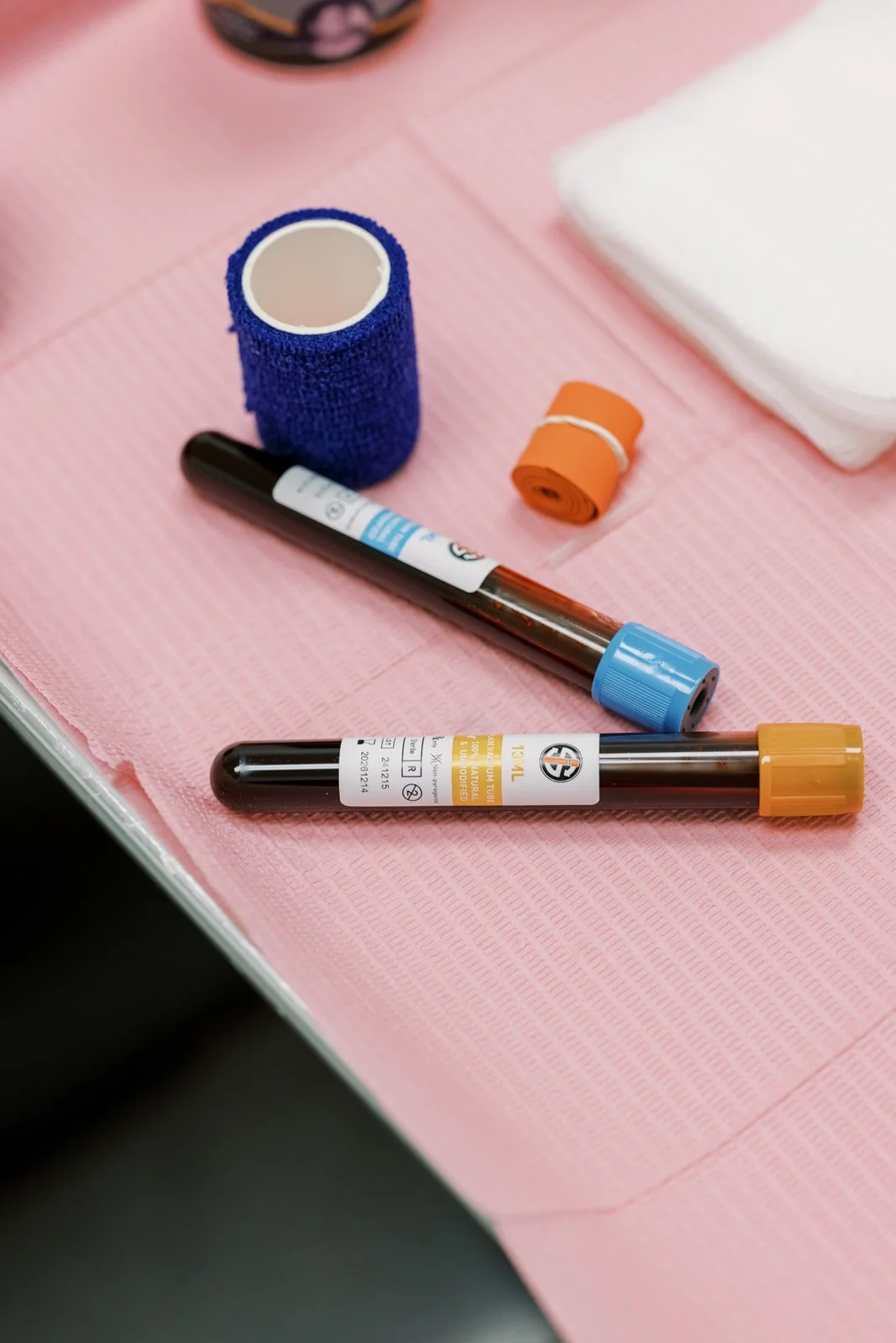 Two blood sample collection tubes with different colored caps, a blue elastic tourniquet, a black marker, and a stack of white gauze pads on a pink medical table.