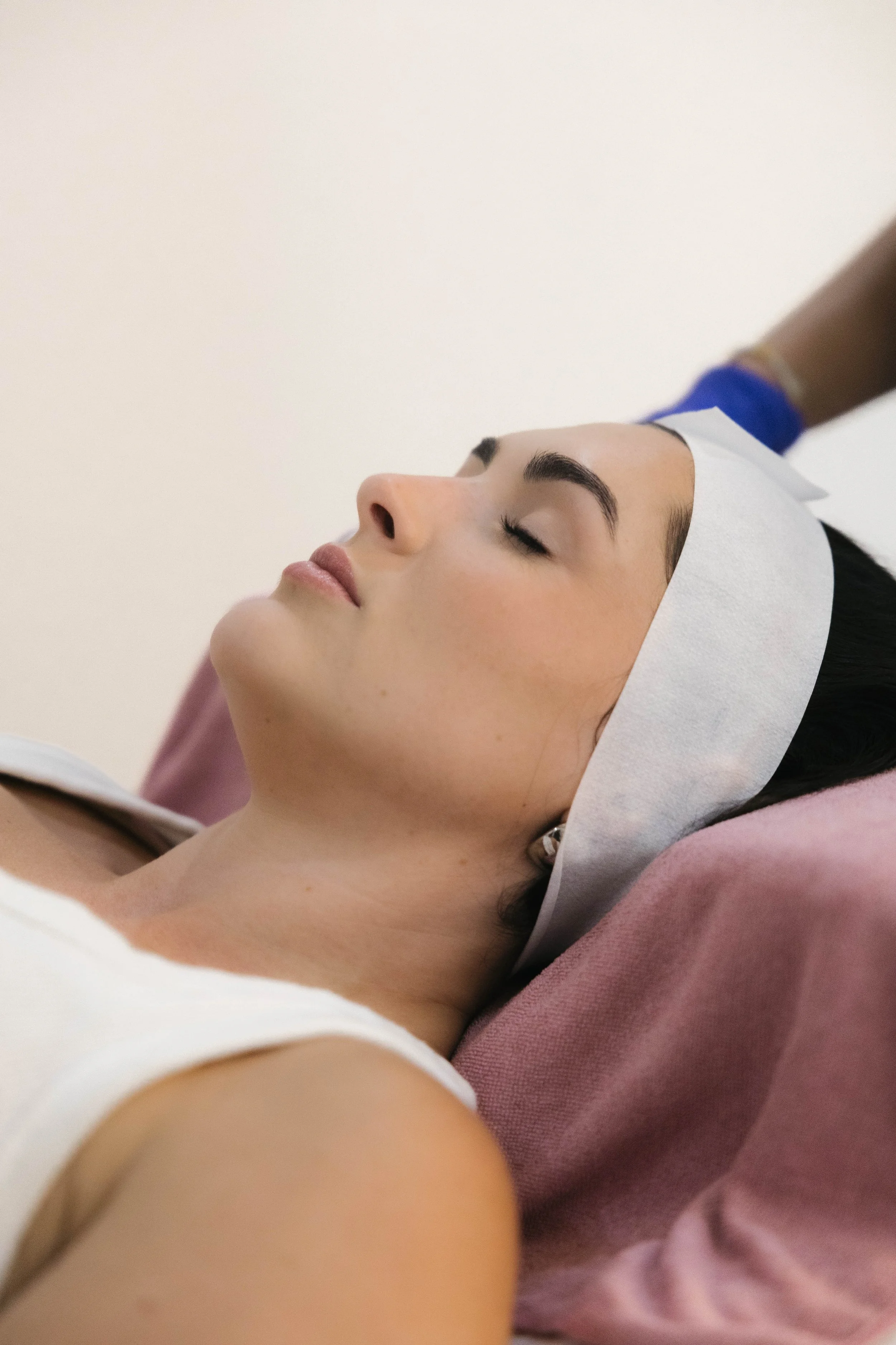 A woman is lying on a black treatment bed with her eyes closed, receiving a facial treatment from a professional in a spa or skincare clinic. The professional is wearing black gloves and a black uniform, applying skincare products to the woman's face. The background shows a cart with skincare supplies including bowls, a sponge, and bottles.