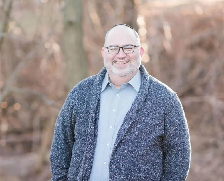 A smiling middle-aged man with glasses and a beard stands outdoors in a natural setting with trees in the background, wearing a light blue shirt and a gray cardigan.