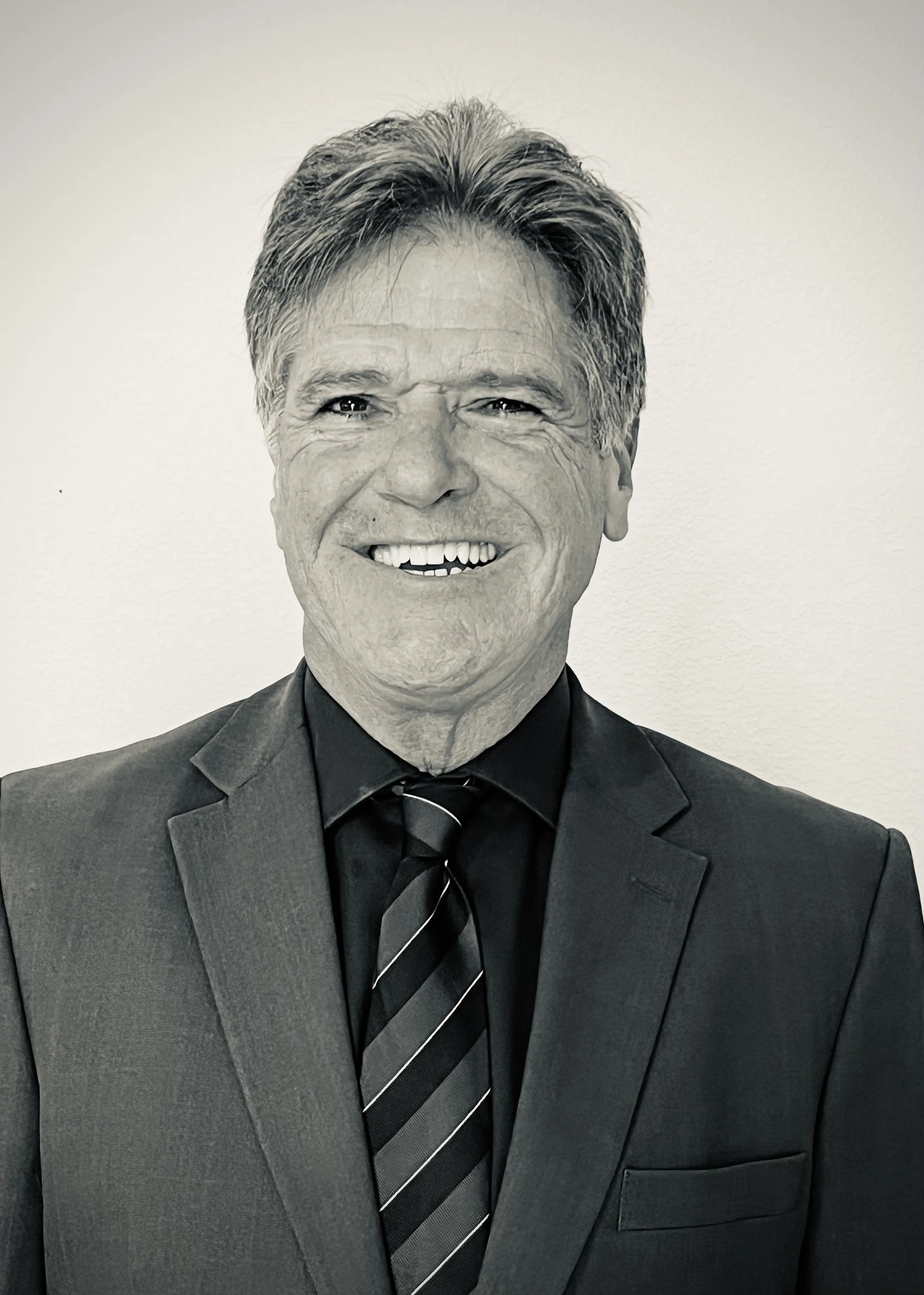 A black-and-white photo of Adam Schultheis, smiling and wearing a suit, tie, and a collared shirt, with a plain light-colored wall in the background.