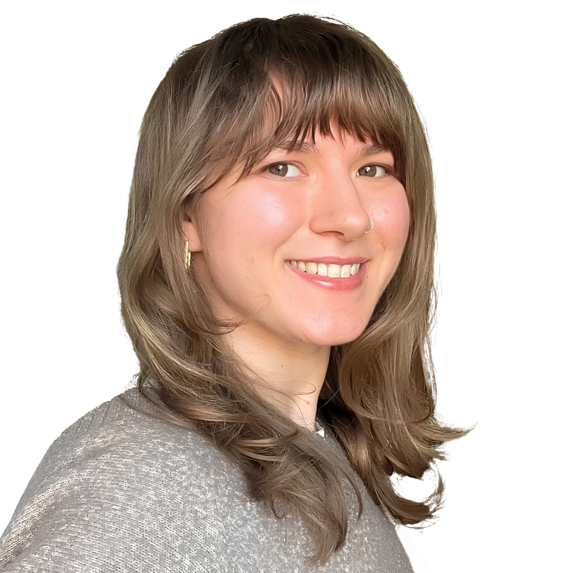 A woman with shoulder-length brown hair and a nose ring, wearing a gray textured top and gold earrings, smiling in front of a white background.