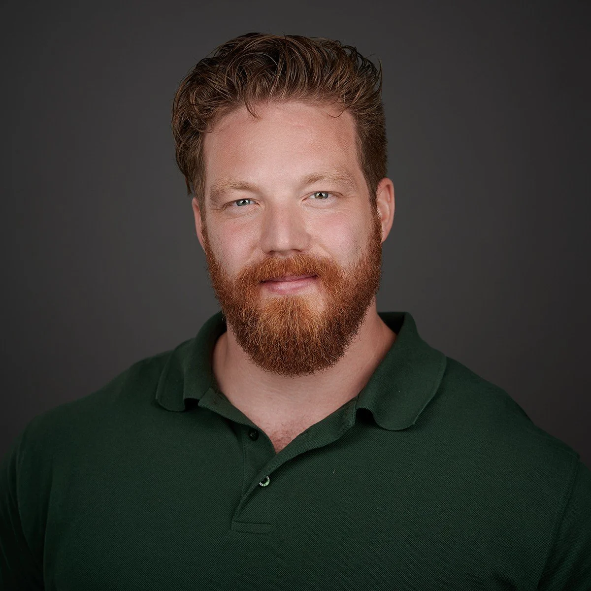 Portret van een man met een rood bebaarde en kort gekapte haardracht, lachend met een groene polo, tegen een grijze achtergrond. Mathijs Labee