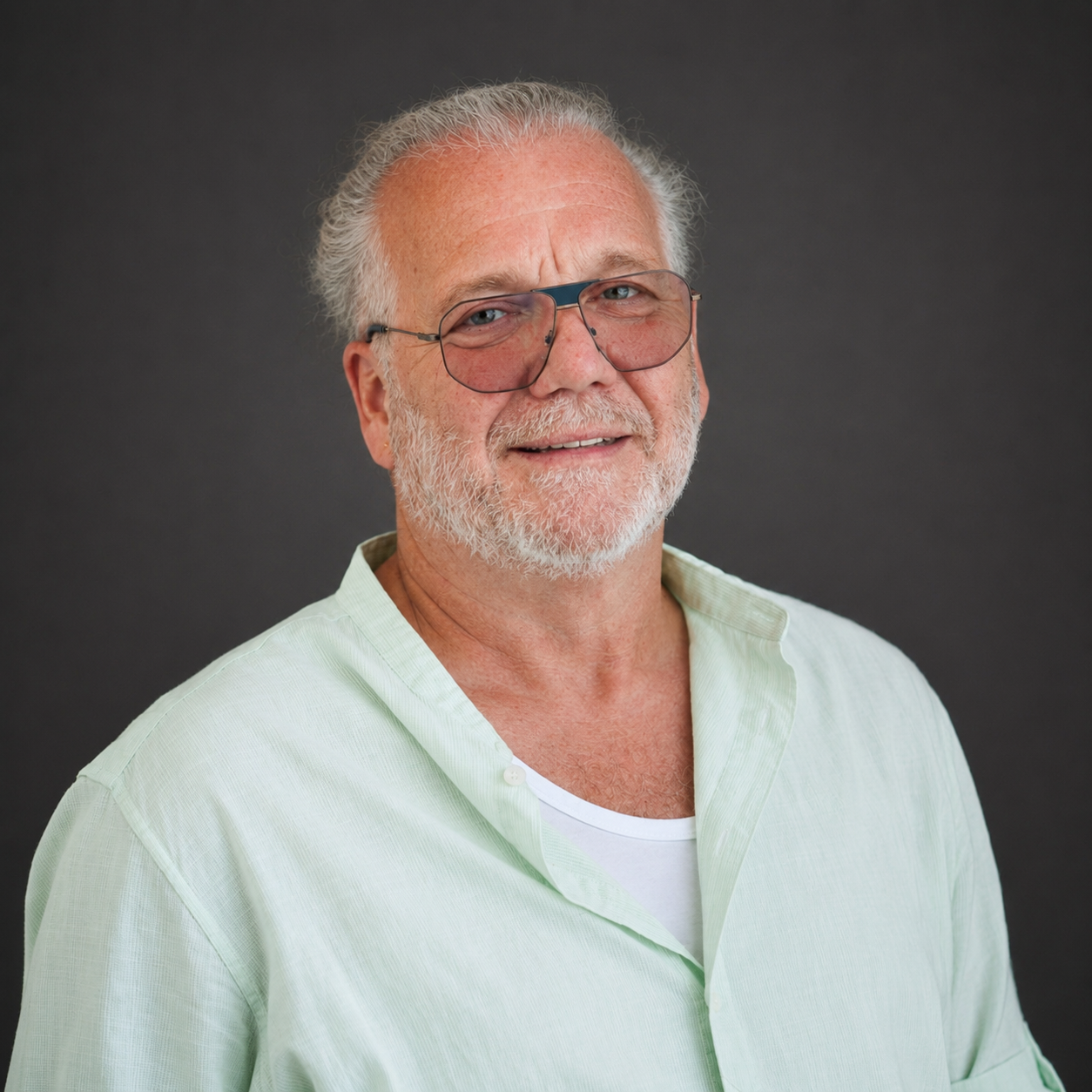 Portret van een oudere man met witte haar en baard, draagt een lichtgroen overhemd en bril, lachend voor een grijze achtergrond. Jan Labee