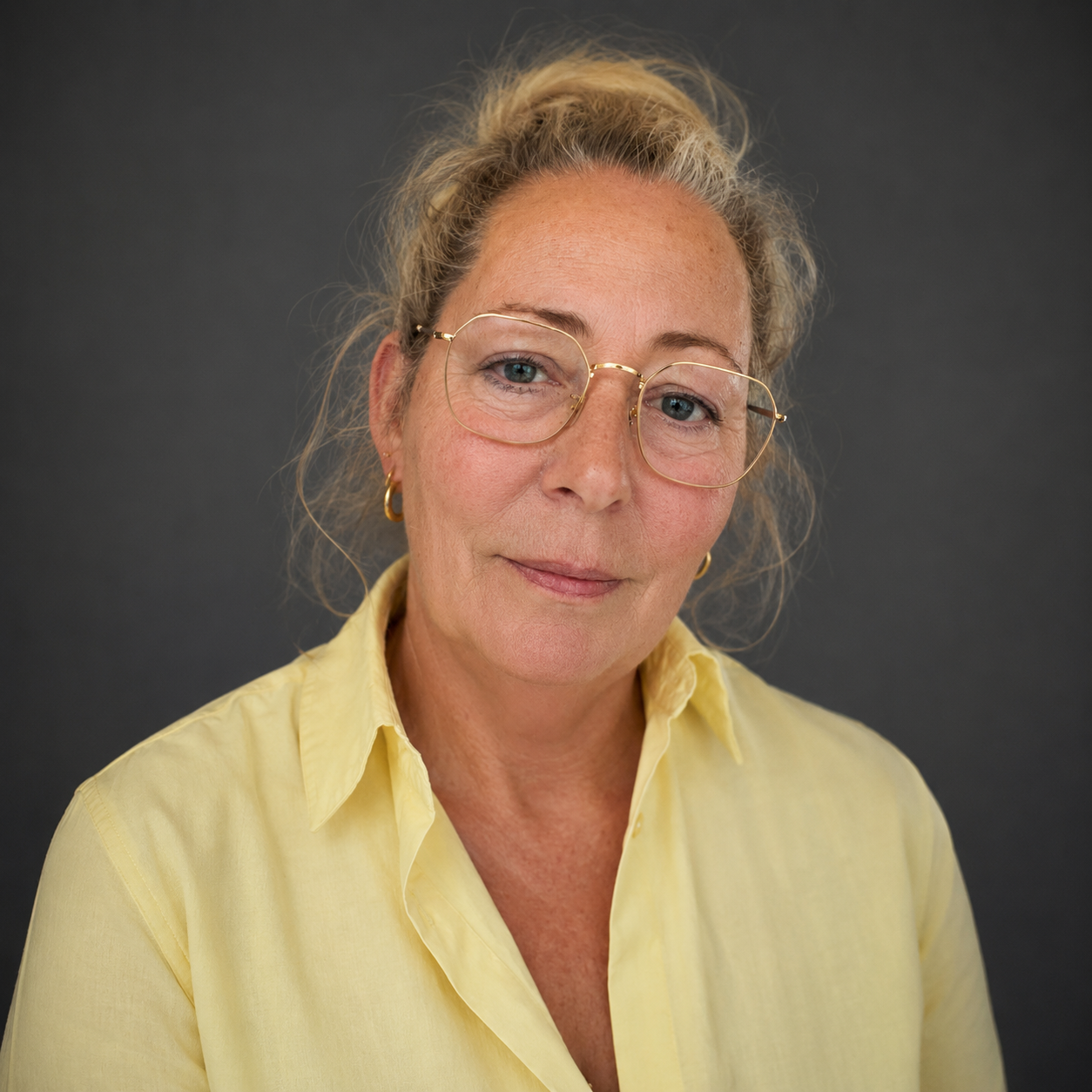 Portret van een oudere vrouw met blond, golvend haar, blauwe ogen, en een vriendelijke glimlach, gekleed in een gele blouse, steekt een geel oorring en ronde bril, tegen een grijze achtergrond.