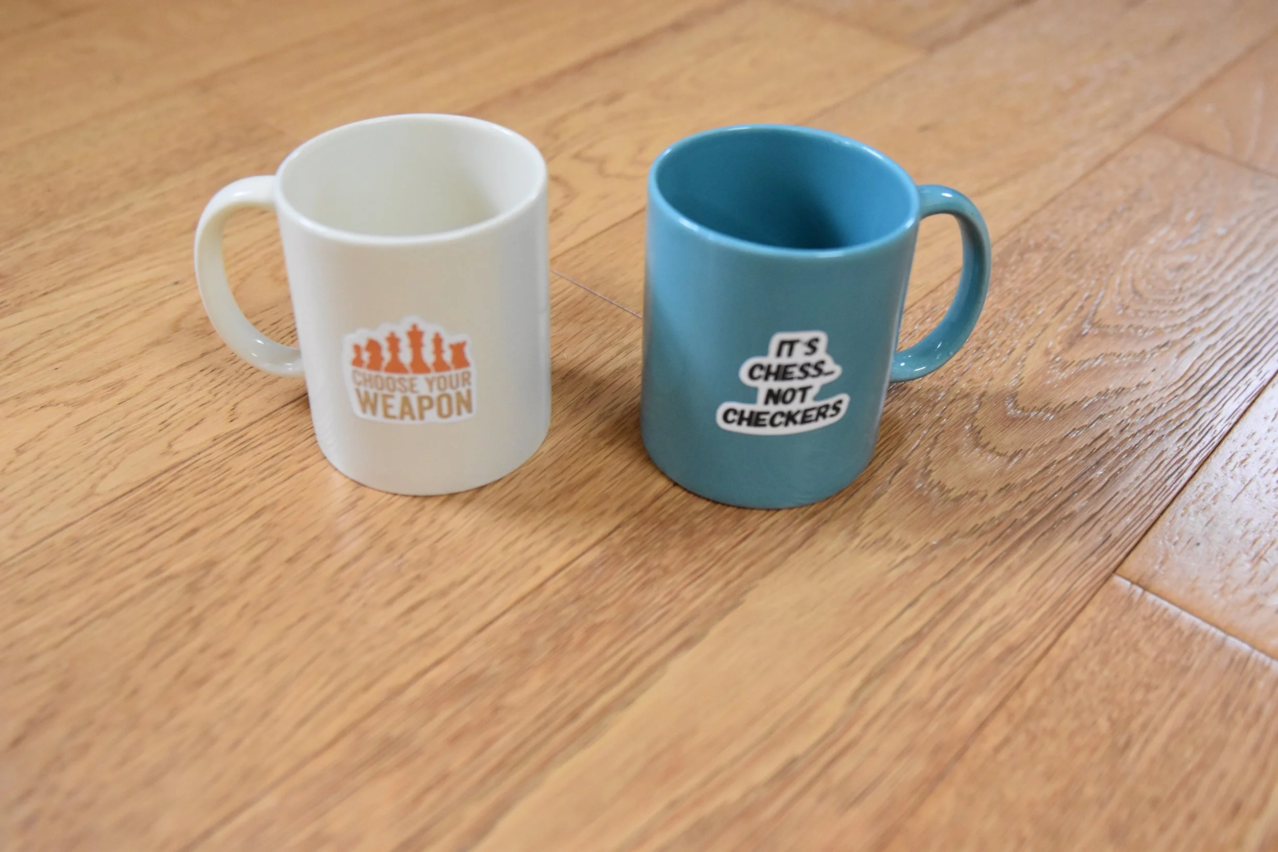 Two coffee mugs on a wooden table, one white with an orange chess piece graphic and text, the other blue with white text.