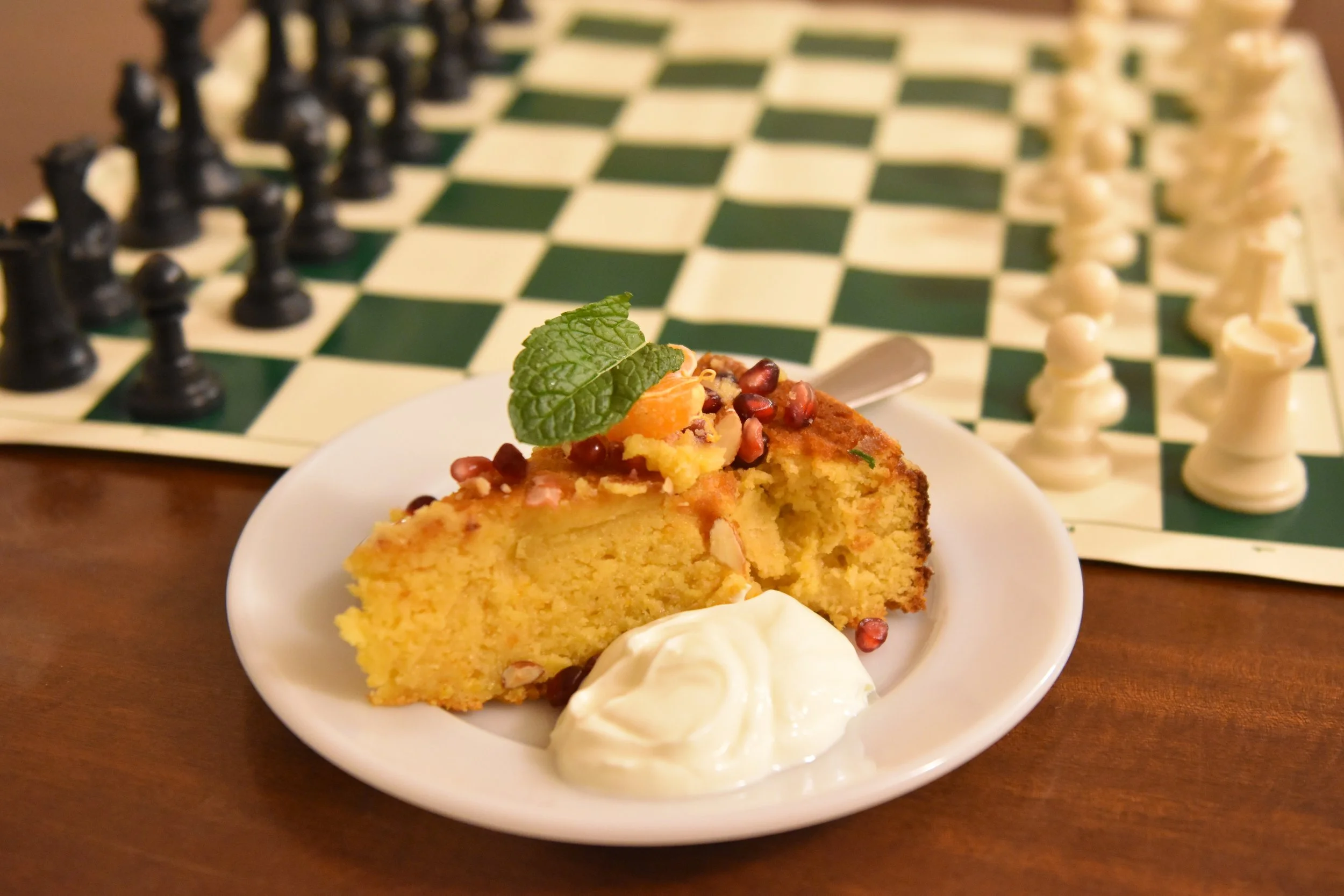 A slice of yellow cake with fruit and nuts on top, garnished with a mint leaf, served with whipped cream and a dollop of cream or ice cream on a white plate. In the background, a chessboard with black and white pieces is visible.