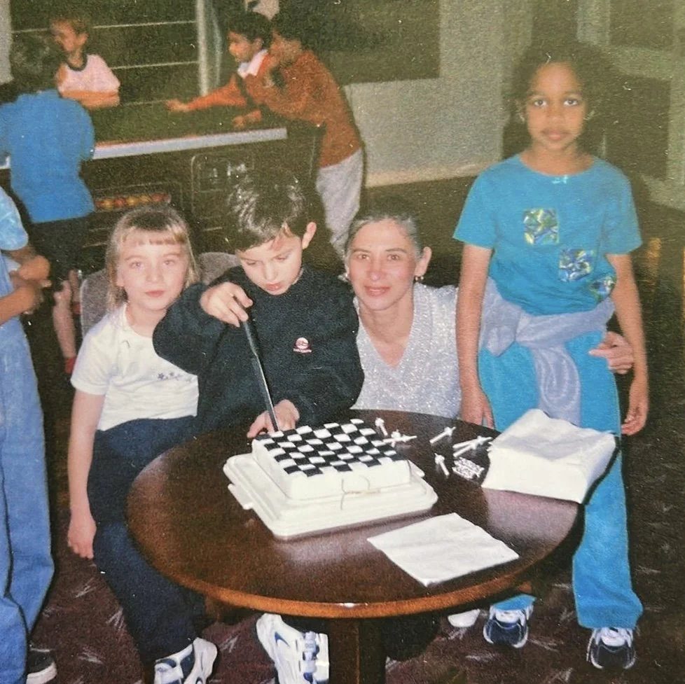 A family celebrating a birthday with a chess-themed cake at a party, children gathered around, one girl standing and others sitting, some in casual clothes, in a room with a table and some kids in the background.