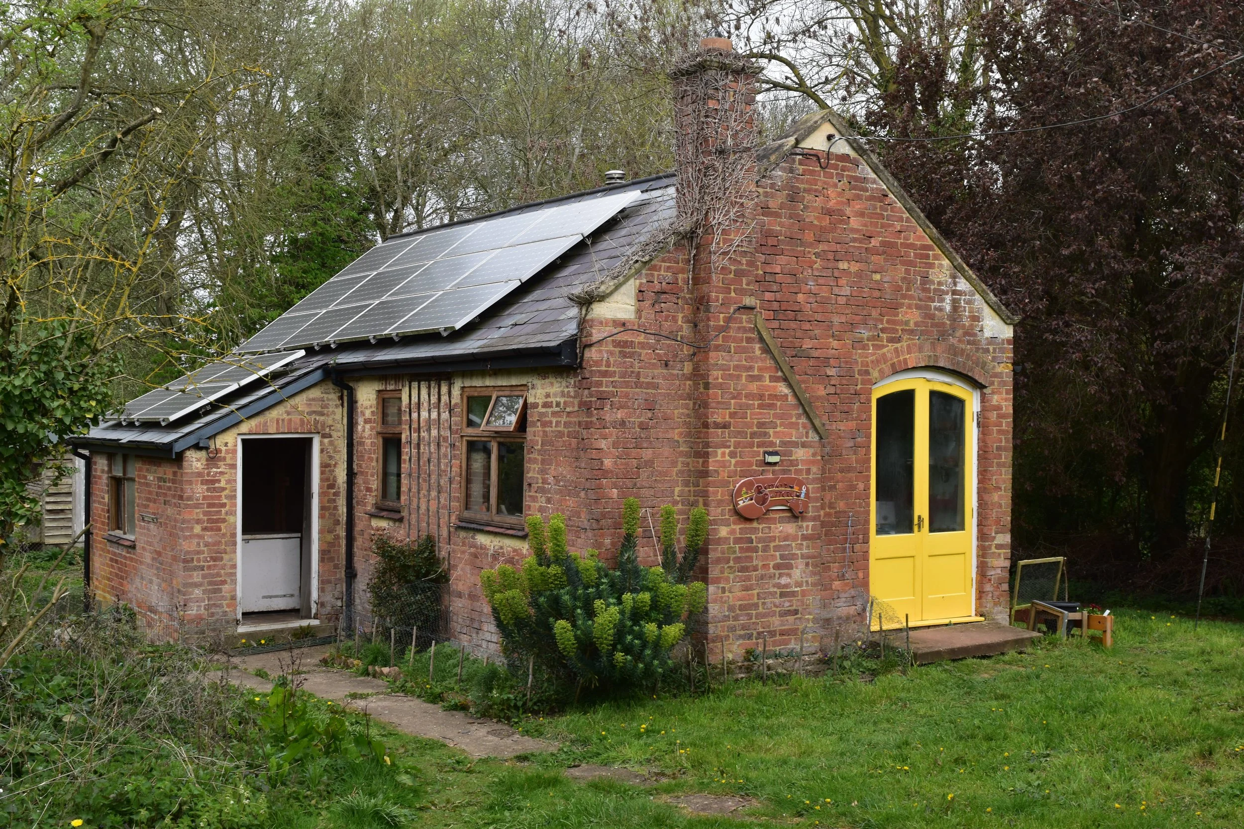 A small brick house with a yellow door, solar panels on the roof, and greenery around it, including trees and bushes.