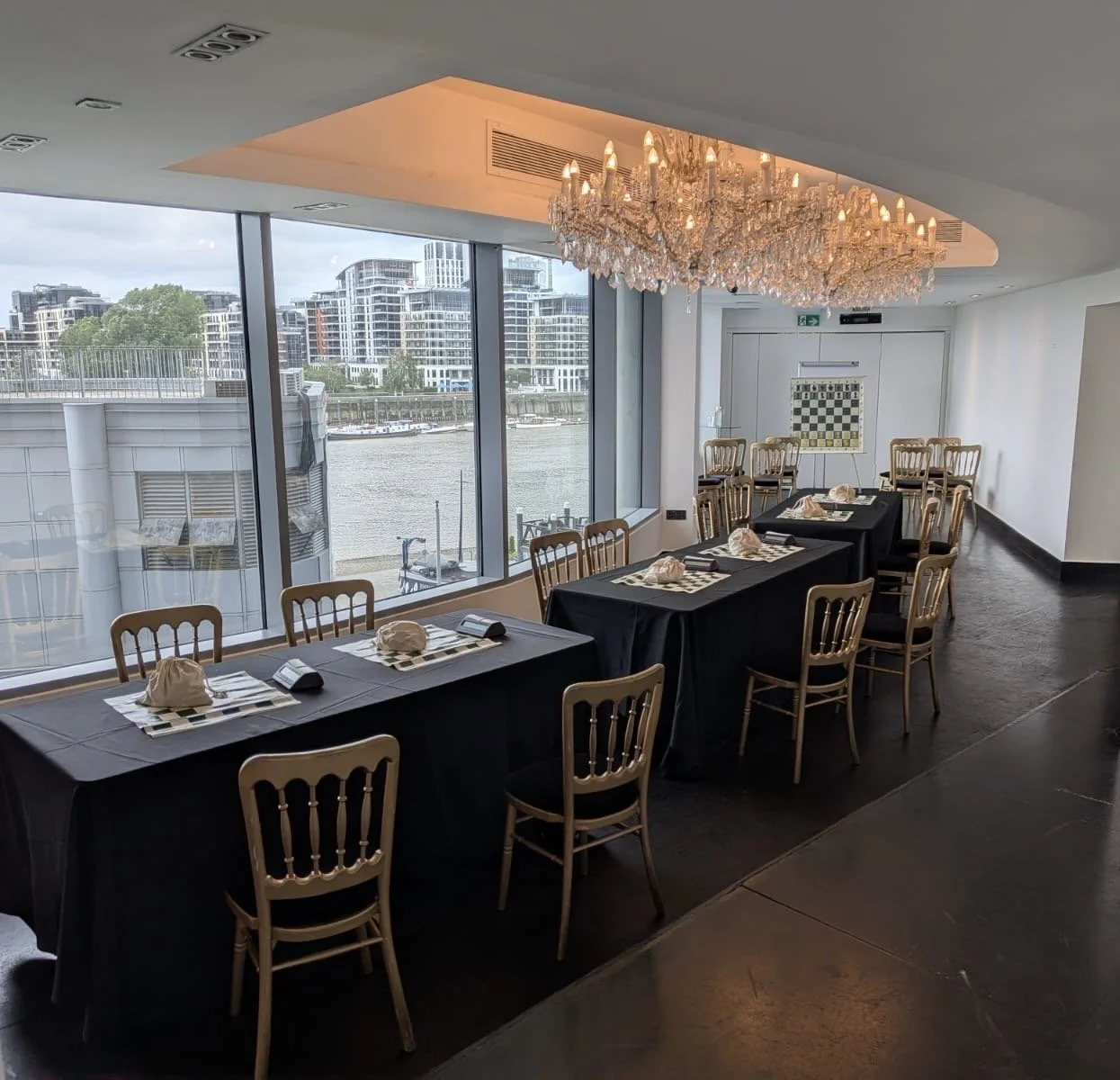 A restaurant or event space with large windows overlooking a river and cityscape, decorated with a chandelier, arranged with tables covered in black tablecloths, set with checkered placemats, napkins, and cloth bags, and surrounded by gold chairs.